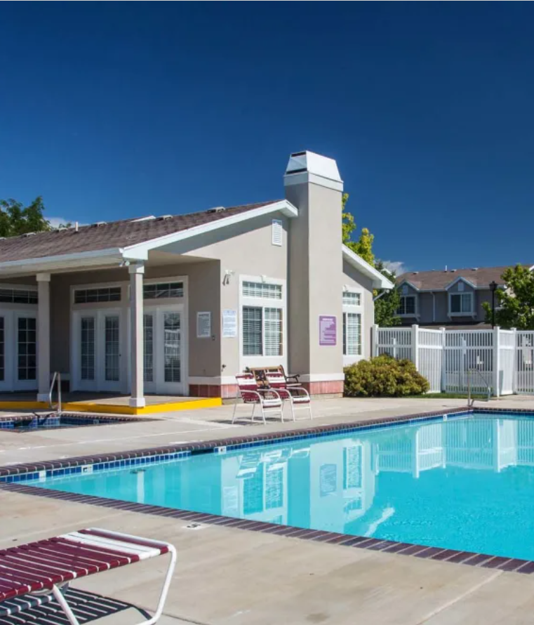View of bright blue swimming pool and resident building with two sets of double French doors, a square hot tub right in front, and one maroon and white lounge chair off to the side
