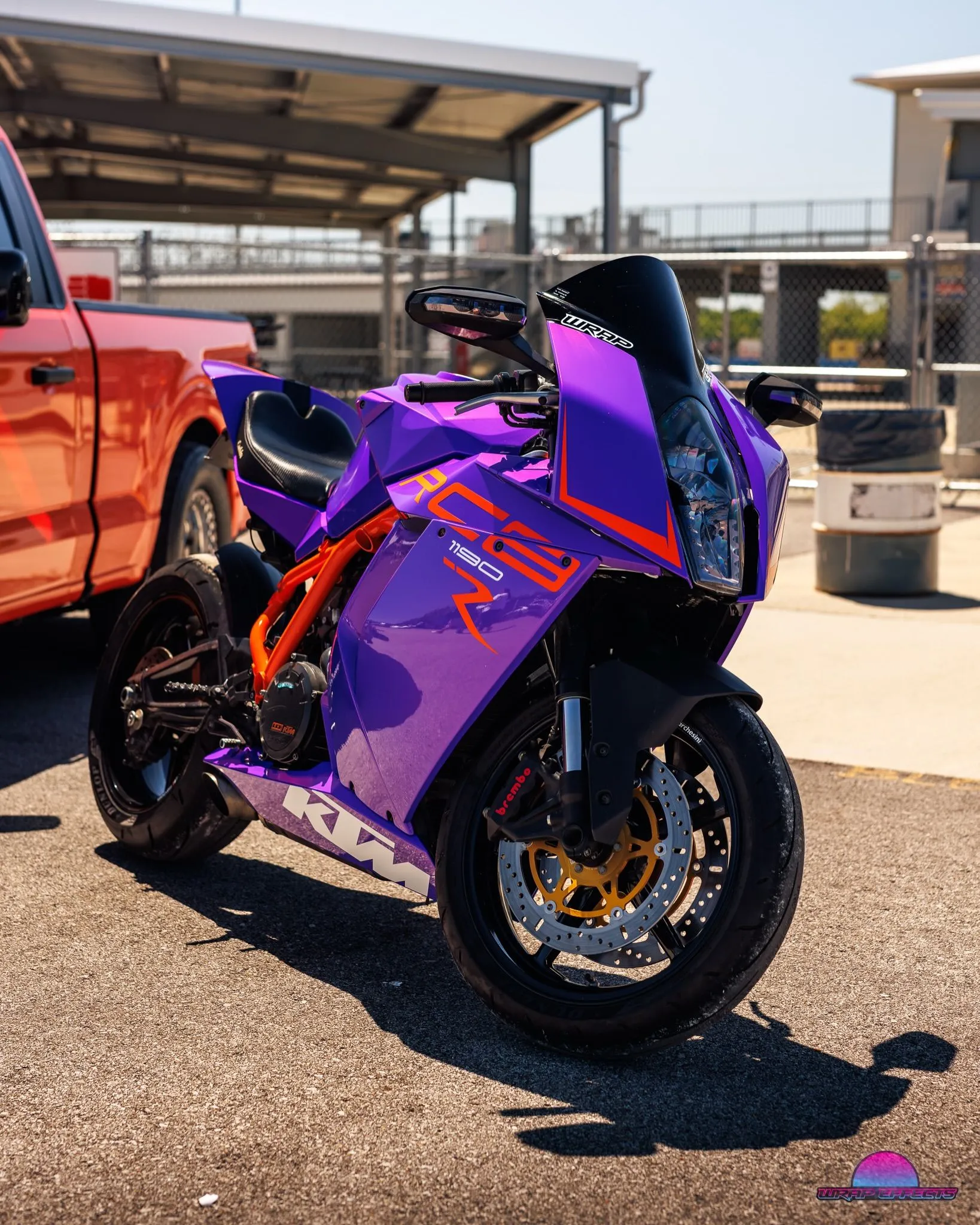 Customized purple KTM RC8 sport motorcycle featuring vibrant graphics, orange frame, premium brakes, aerodynamic fairings, and professional wrap design displayed outdoors under bright sunny conditions near paddock.