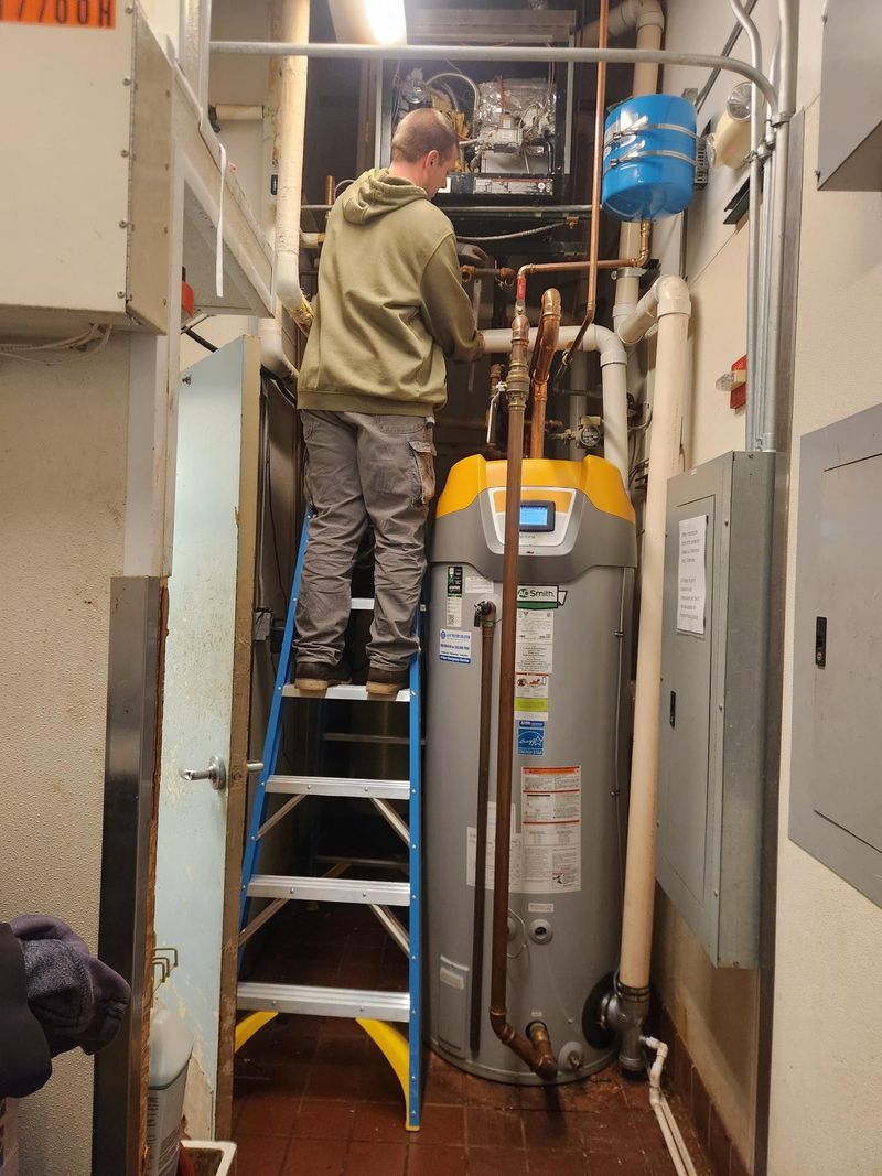 A professional technician stands on a blue ladder to perform maintenance on a tall, high-efficiency A.O. Smith Cyclone commercial water heater, surrounded by intricate copper piping and overhead mechanical components.
