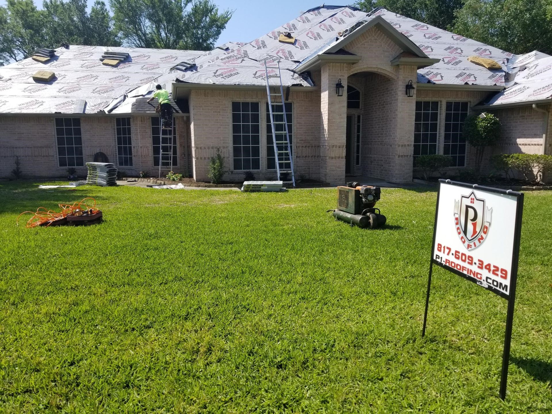 Roof replacement is underway on a single-story brick residence with underlayment installed, ladders positioned, and roofing materials prepared for the next phase of installation by professionals.
