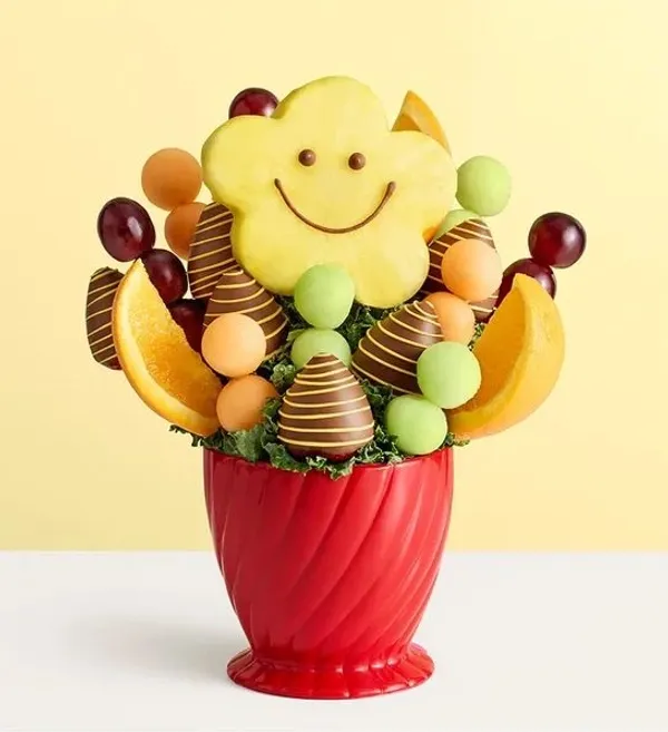 A vibrant fruit bouquet shaped like a smiling flower is presented in a swirled red vase, featuring c 