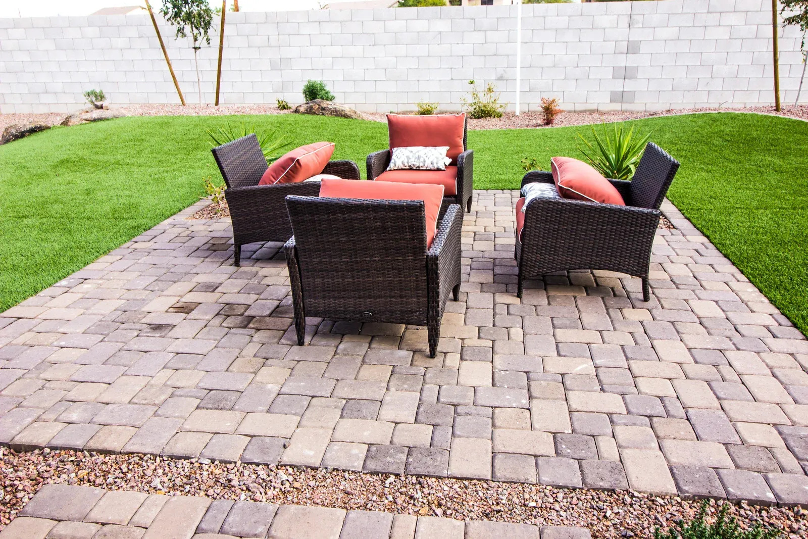 A cozy outdoor living space features a paved patio with four dark wicker chairs and orange cushions arranged around a central spot, surrounded by green grass and a block wall.