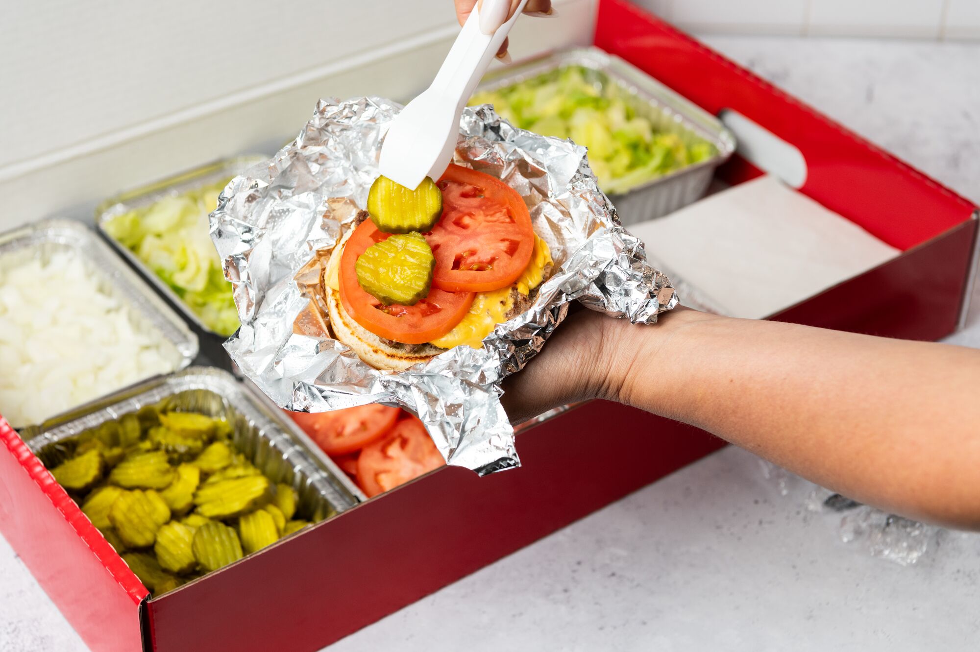 A close-up photo of a person putting pickles on their cheeseburger in front of a Five Guys catering  Five Guys Thousand Oaks (805)496-0173