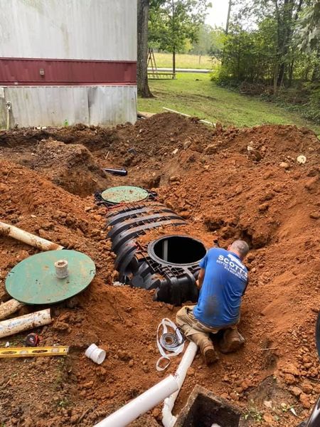 A service technician in a blue Scott's Septic Tank Service shirt installs a large, black plastic septic tank in a deep excavation of red clay soil near a residential building.