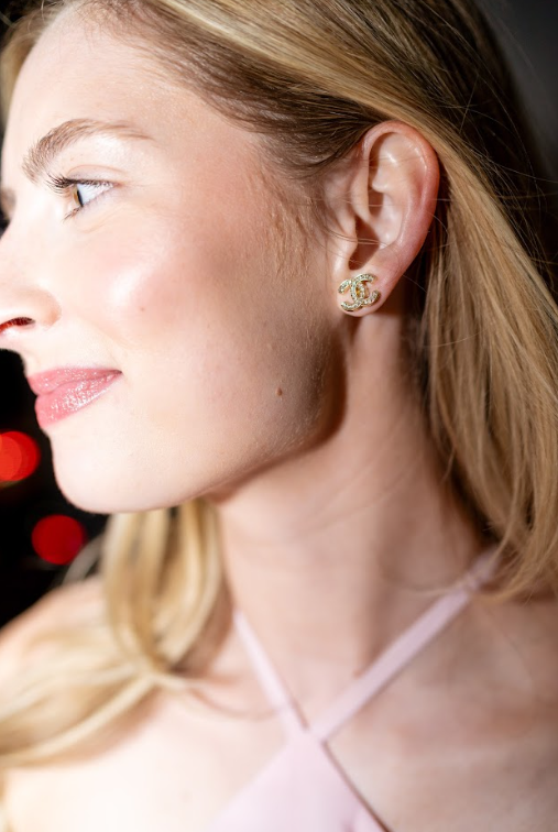A gold-toned interlocking logo stud earring with sparkling accents complements a pastel pink halter top. Discover elegant earrings and curated accessories at Eliana Curated in Old Town Alexandria, VA.