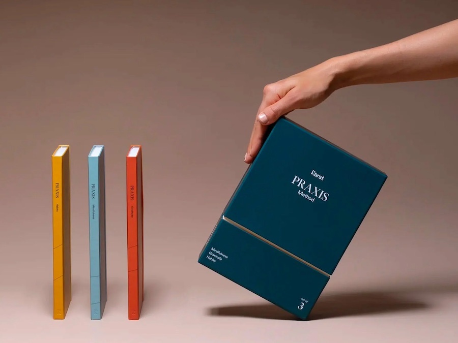 A professional product visual by Tabula Rasa Social Stationers, highlighting the Karst Praxis Method 3-Journal Set. The image features a sleek, dark teal box, labeled "Karst PRAXIS Method" for Mindfulness, Gratitude, and Habits, being presented by a hand. Standing beside it are three slim, vibrantly colored Karst stone paper journals in yellow, light blue, and red. This eco-friendly set, crafted from sustainably sourced recycled stone, offers a silky-smooth, tear-resistant, and waterproof writing experience, embodying a groundbreaking blend of sustainability and a refined journaling practice.