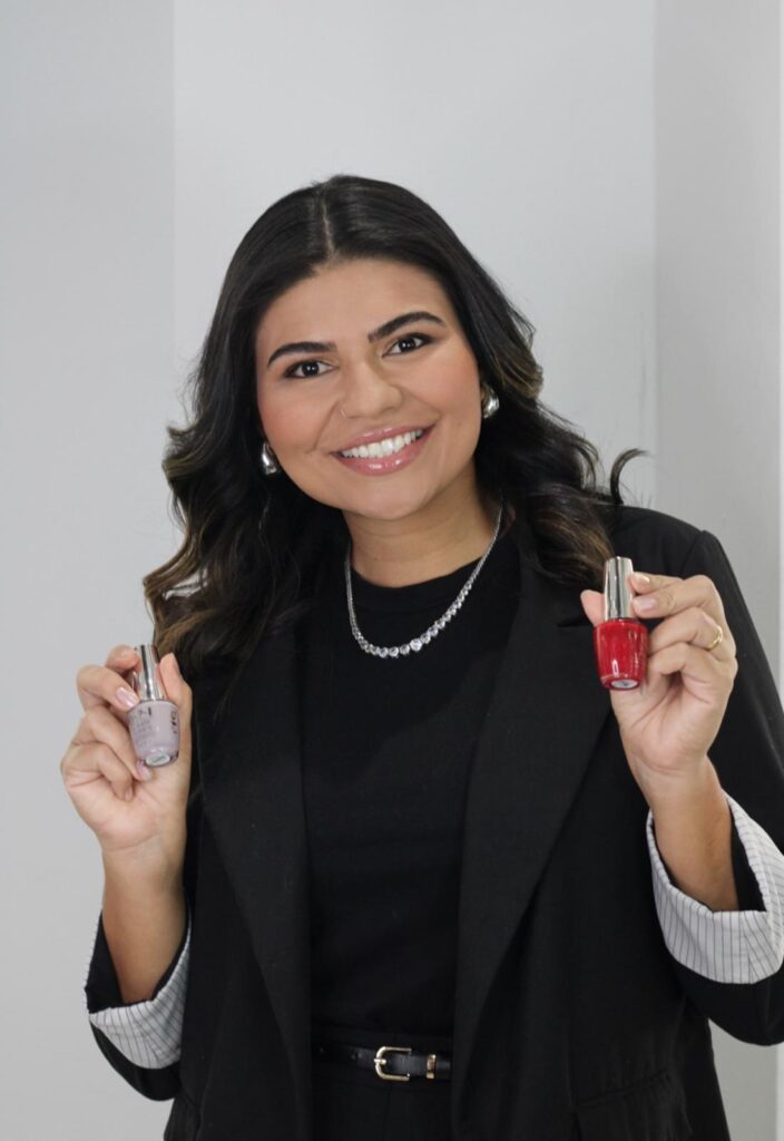 A smiling woman with dark wavy hair, wearing a black blazer over a black top and a sparkly necklace, holds up two bottles of nail polish, one red and one a light pastel color, in front of a plain white background.