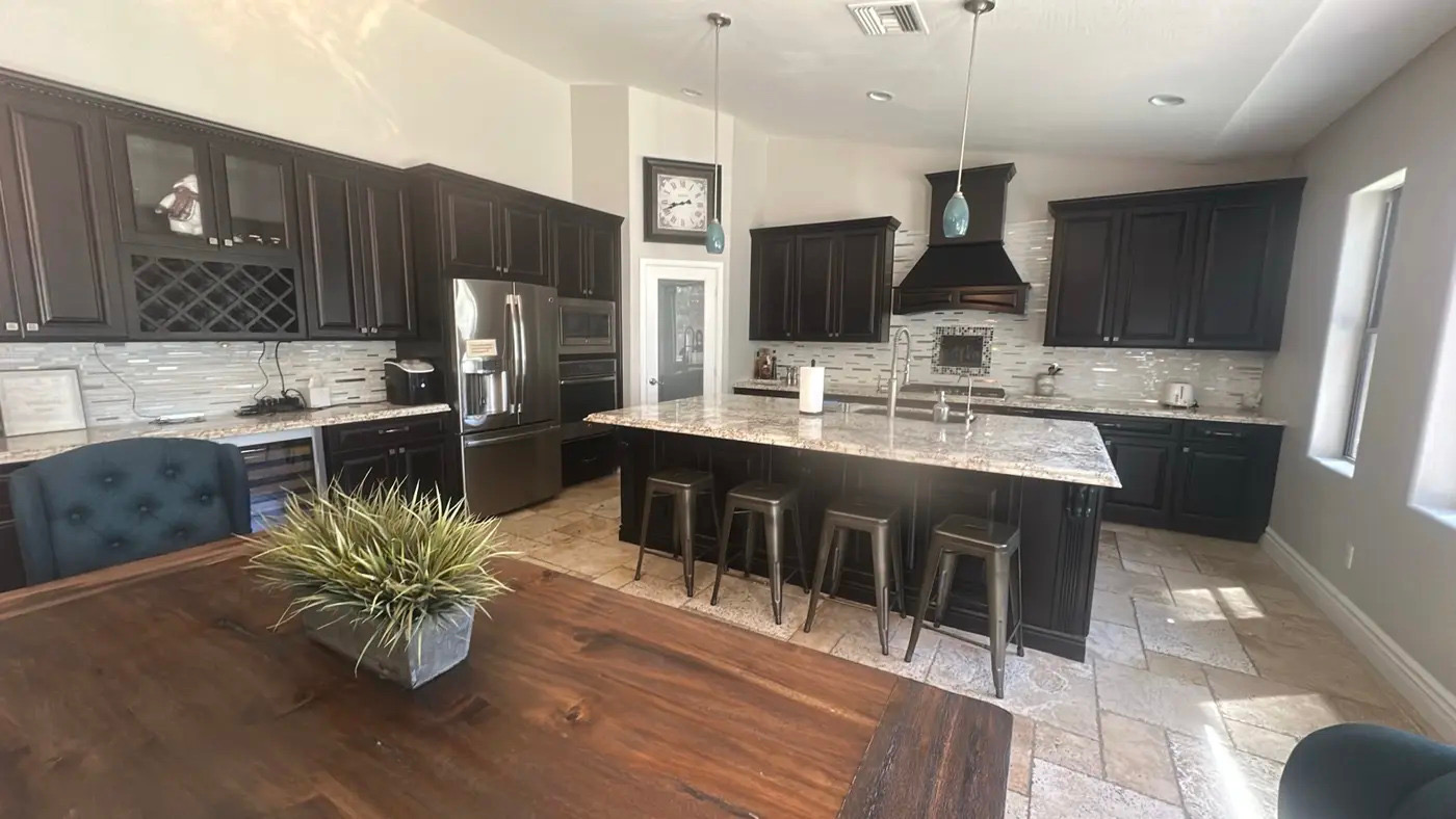 A spacious, modern kitchen with dark wood cabinets, granite countertops, and stainless steel appliances is captured in a wide-angle shot. A large island with seating is positioned in the center, and a dining table with a plant is in the foreground. The overall impression is one of a clean and well-appointed home.