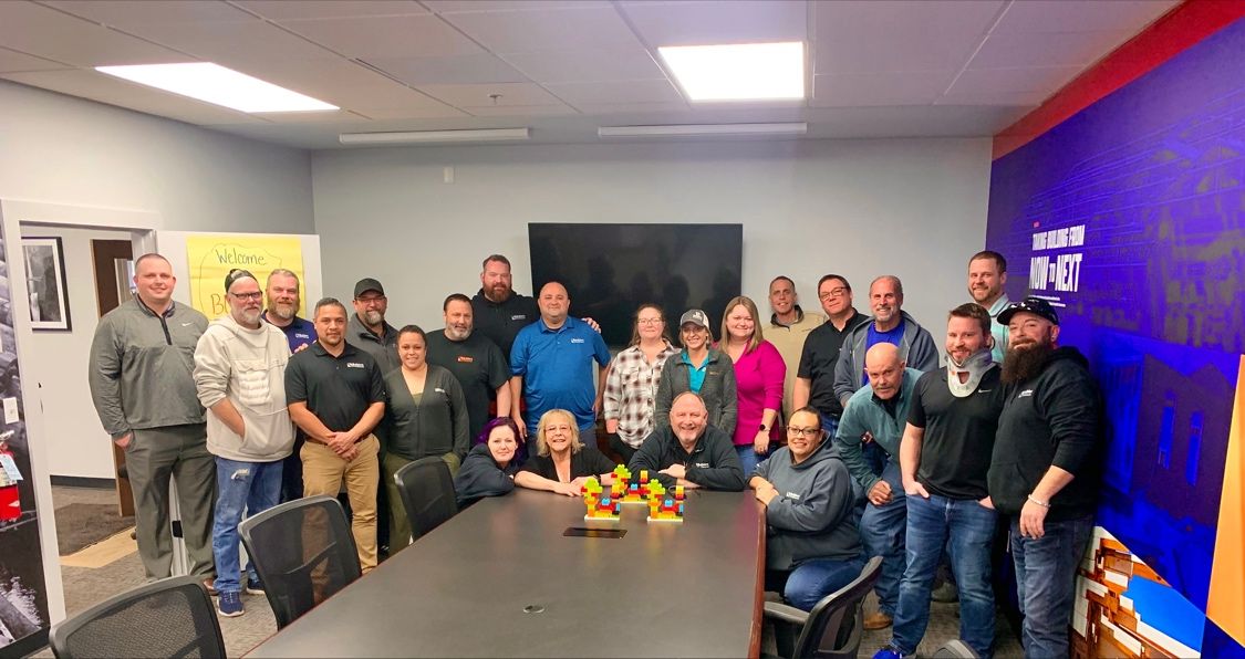 A group of people gathered in a conference room, smiling for a group photo. The room features a large table with colorful building blocks at the center, and a bold wall graphic with the phrase 'Moving from Now to Next' in the background, reflecting a focus on leadership and company values.