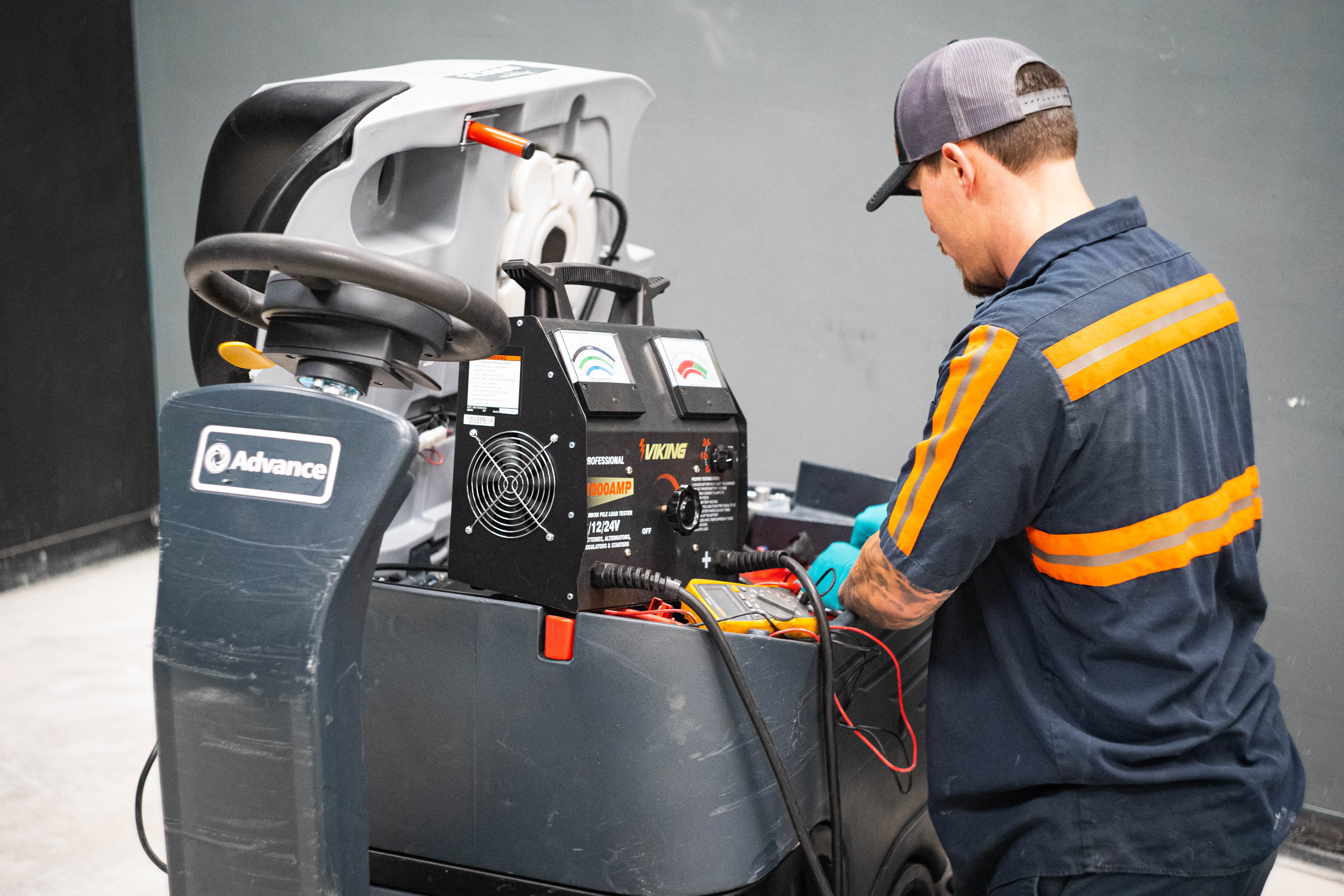 A rear-view action shot of a WaySolutions technician conducting a battery system health check on an Advance commercial ride-on floor scrubber. The technician is operating a black Viking professional 1000-amp 12/24V carbon pile load tester equipped with dual analog diagnostic meters, checking the industrial battery banks for voltage drop and load capacity.