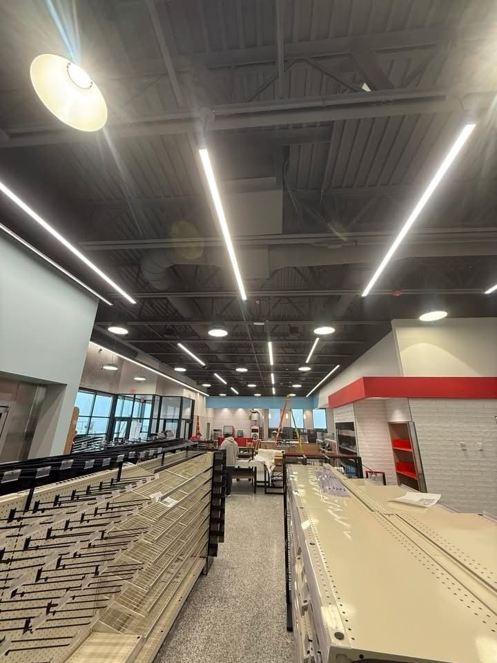 Retail store interior under construction with empty shelving units, exposed ceiling lighting, and work equipment, showing installation progress and preparation for product displays in commercial environment.