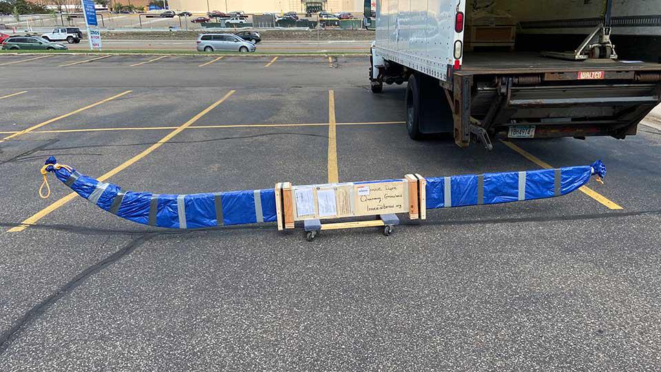 A long wooden crate the shape of curved chopsticks is wrapped in moving blankets, ready to be loaded into the truck in the background.