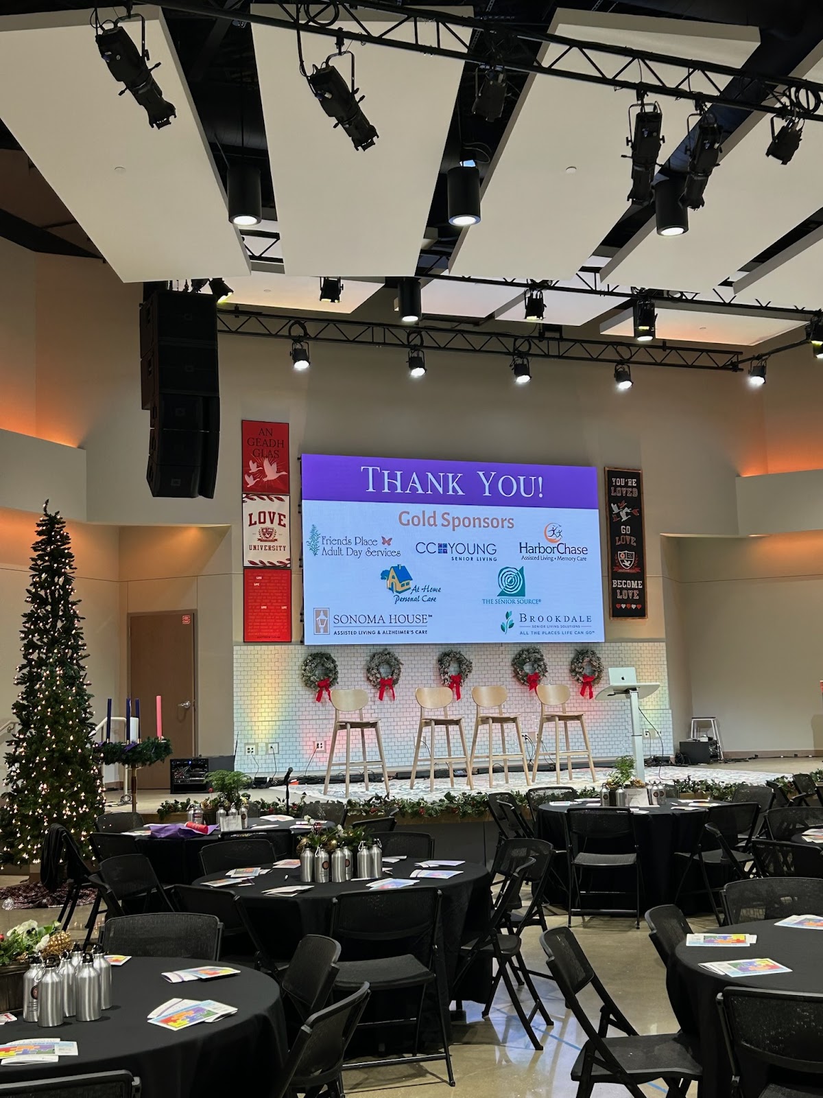 An event stage set up for a ceremony or presentation, with a large screen displaying 'THANK YOU! Gold Sponsors'. Several logos of sponsoring companies are visible on the screen. In front of the screen, there are five stools, and a stage decorated with wreaths. To the left of the stage, a decorated Christmas tree stands next to a display of four advent candles. The foreground shows round tables draped with black tablecloths, set up for attendees with water bottles and pamphlets. The overall setting appears to be an indoor venue, possibly a conference hall or auditorium.