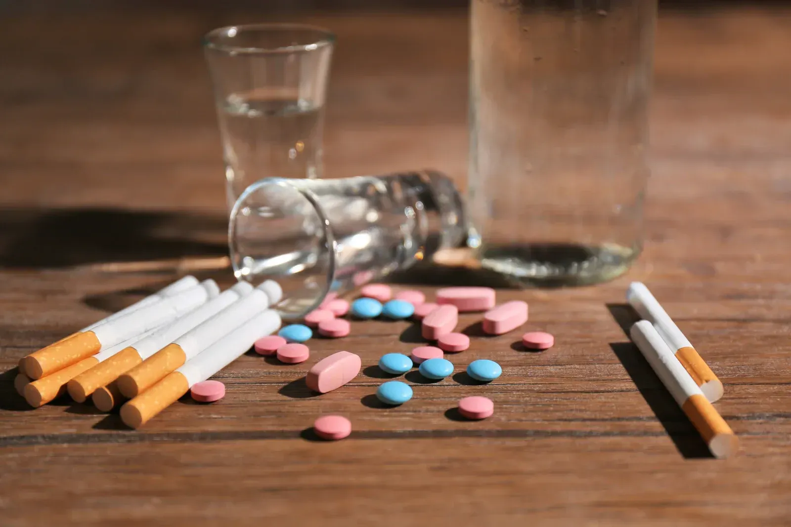 The image displays cigarettes, various pink and blue pills scattered on a wooden surface, and two empty glasses, one lying on its side. A partially visible clear bottle stands in the background. This arrangement suggests substance abuse.
