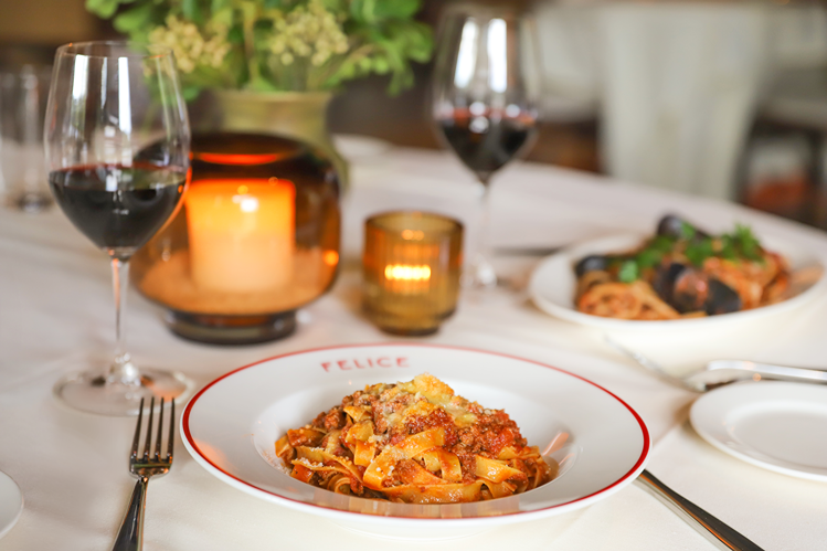 A close-up of a hearty pasta dish on a white tablecloth, accompanied by two glasses of red wine and glowing candles. The soft lighting creates an intimate dining experience.