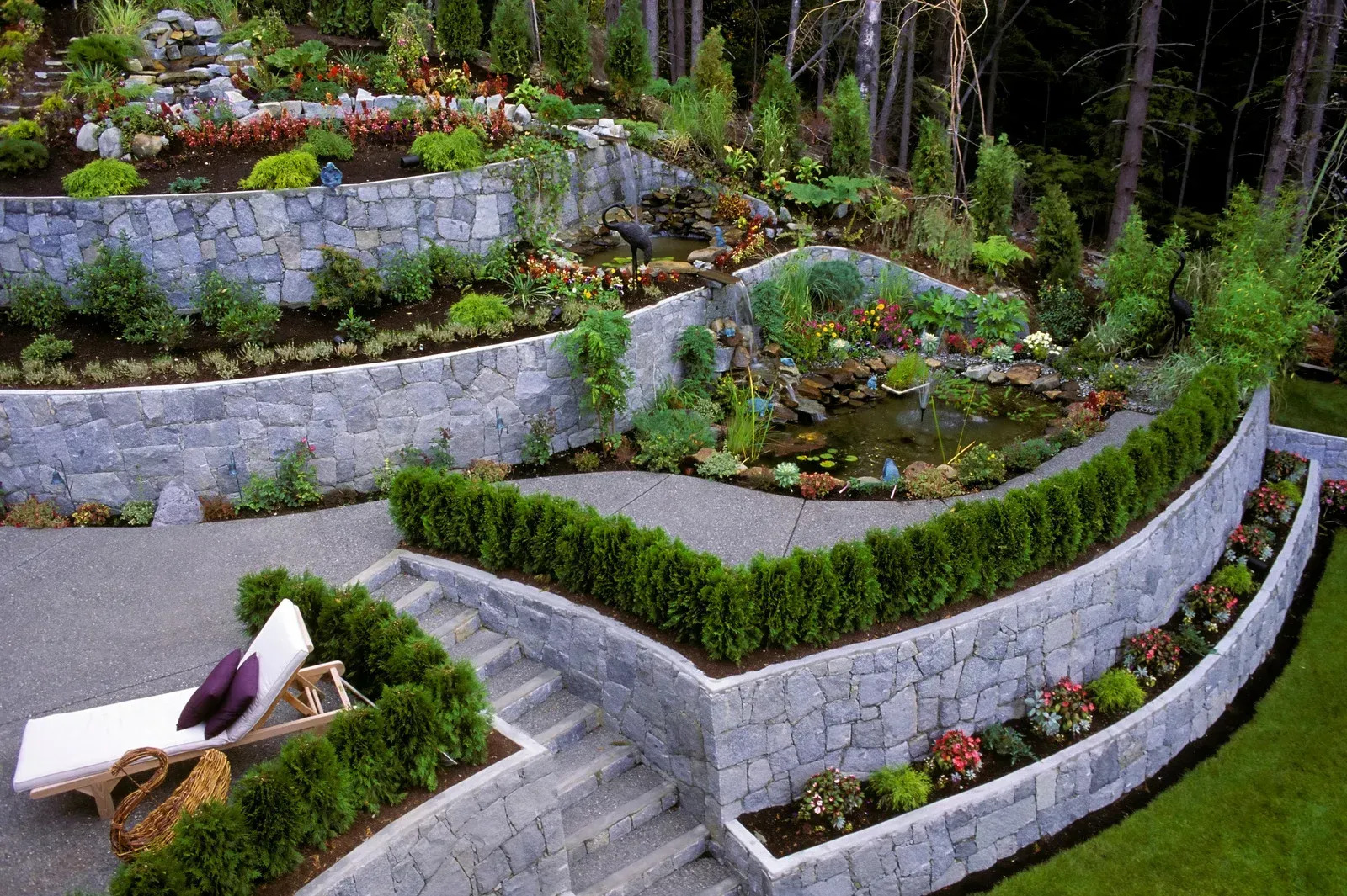 An elaborate terraced garden features stone retaining walls, lush plantings, steps, a pond, and a lounge chair, creating a serene and beautifully landscaped outdoor space.