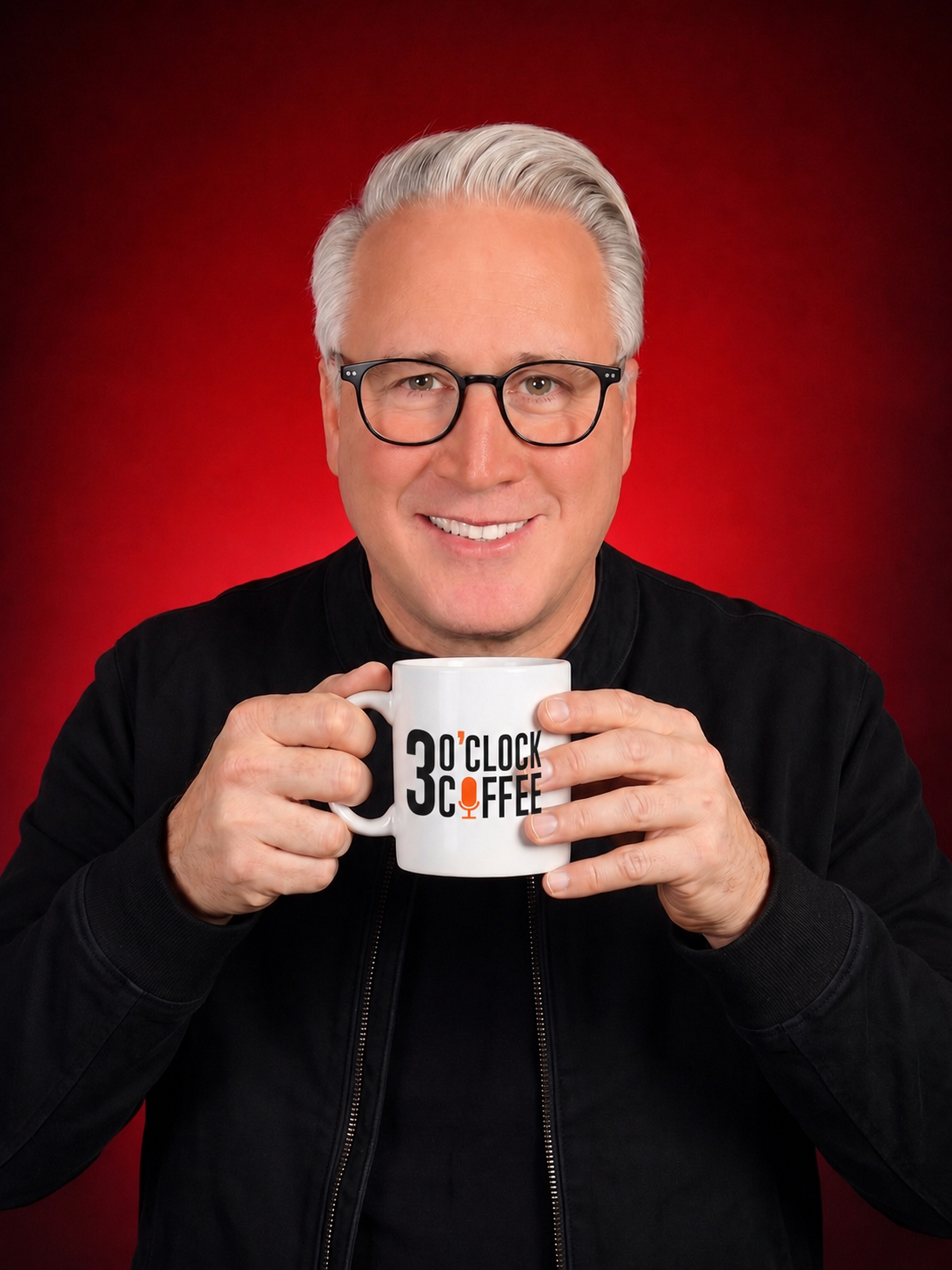 A man with gray hair and glasses smiles while holding a white mug with black and orange text and a microphone graphic. He is wearing a black jacket and is set against a dark red background.