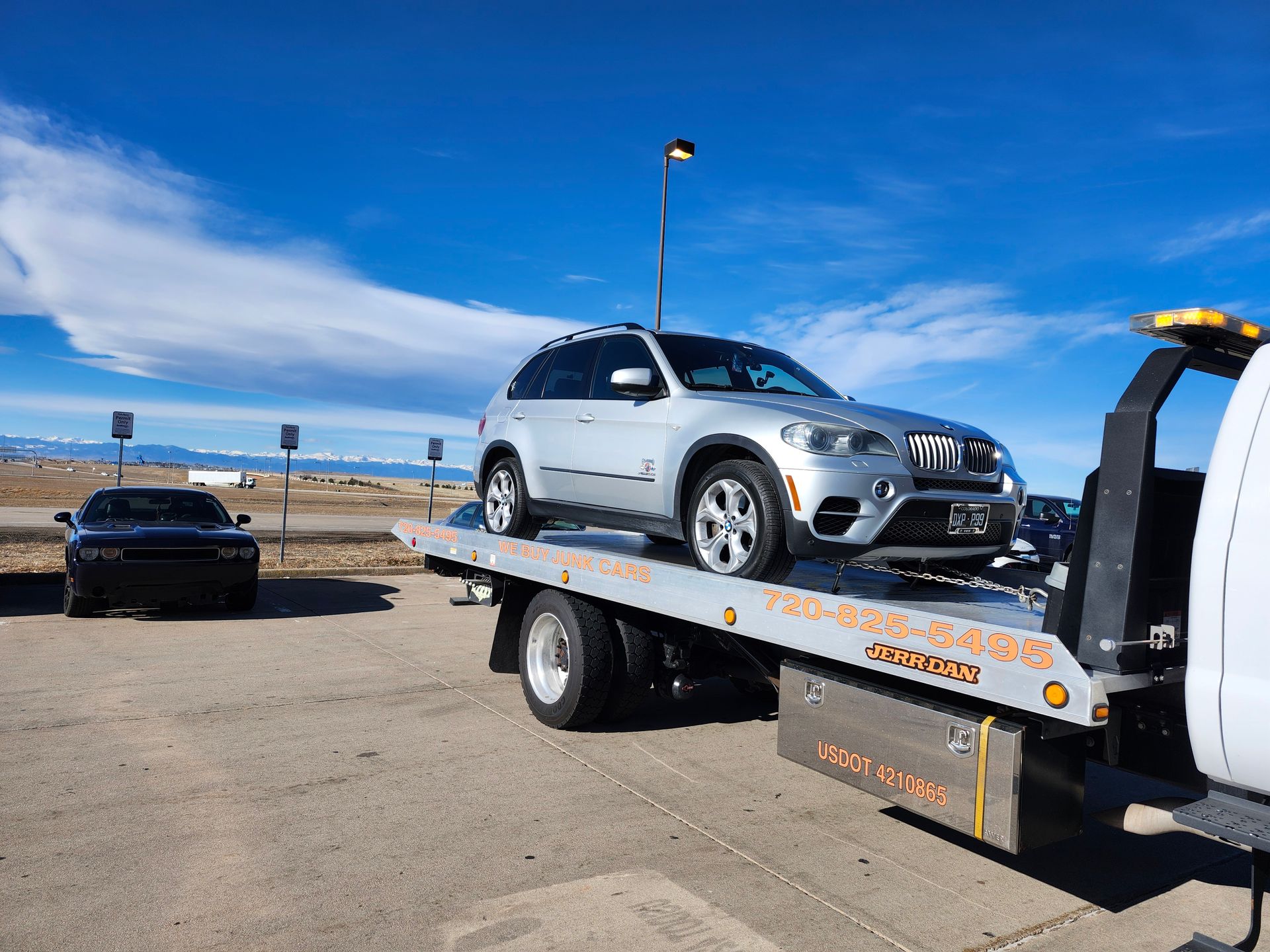 A tow truck carries a white SUV on a flatbed in an open outdoor area, highlighting professional towing services with mountains in the background and clear blue skies.