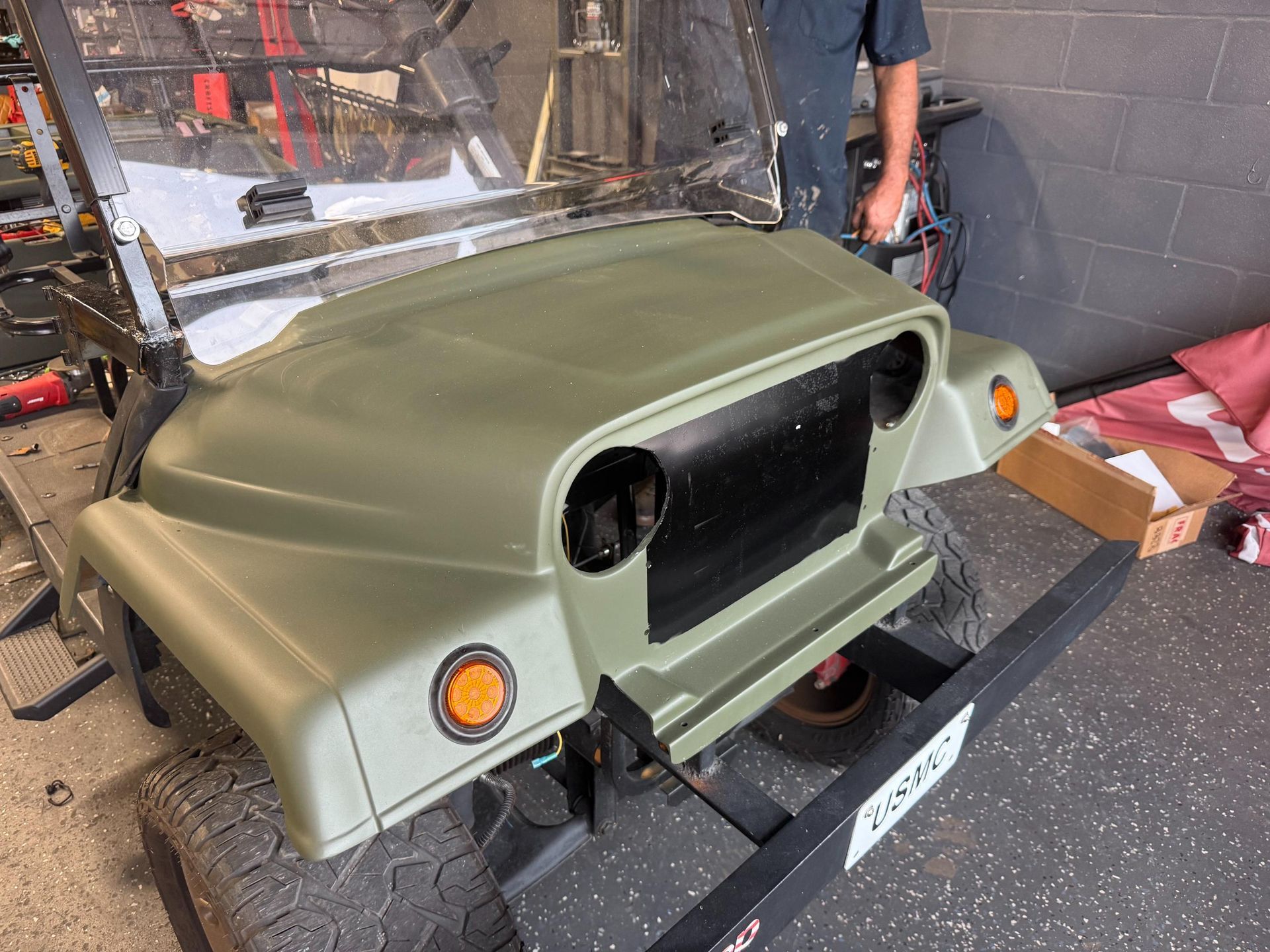 A rugged, custom utility vehicle or golf cart painted in matte olive green is shown in a garage bay. The front end is being worked on, featuring large tires and a "USMC" license plate frame on the bumper.