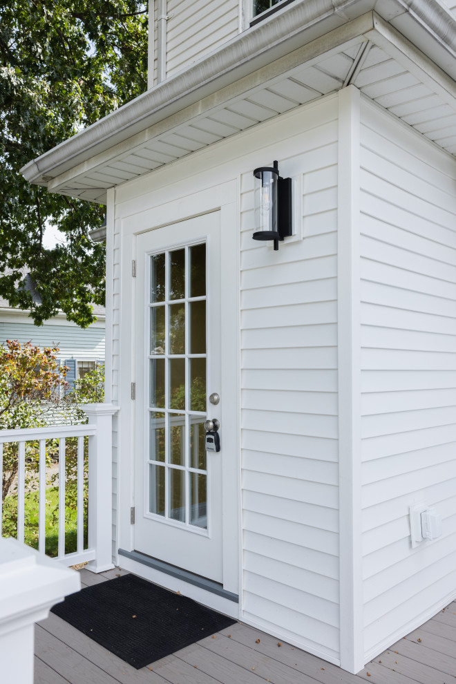 A white paneled door with a glass window and a black outdoor lantern is visible on the side of a white house. A grey deck with a white railing is in the foreground, and a black doormat is on the deck in front of the door. Green trees and a blue house are visible in the background.