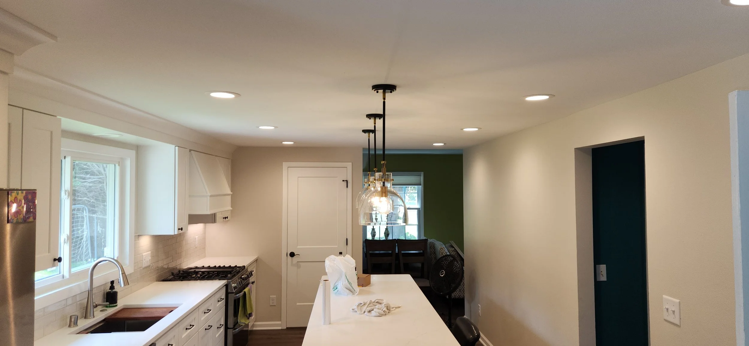 A modern kitchen with white cabinets and countertops. The kitchen features recessed lighting and a set of pendant lights over the island. A gas range is visible on the left, and a sink with a faucet is also present. In the background, there is a white door and a hallway leading to another room with green walls and furniture. A fan is placed near a sofa.