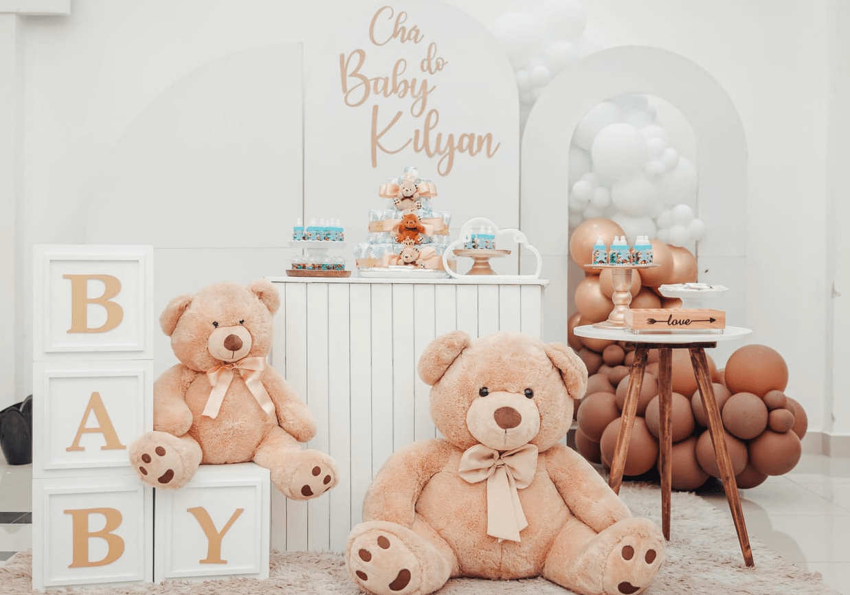 A festive baby shower setup with two large teddy bears, a multi-tiered diaper cake, decorative blocks spelling 'BABY', and balloons. The backdrop features a large archway with gold lettering that reads 'Cha do Kilyan', suggesting a Portuguese baby shower. The overall color scheme is soft and pastel, with accents of gold and beige.
