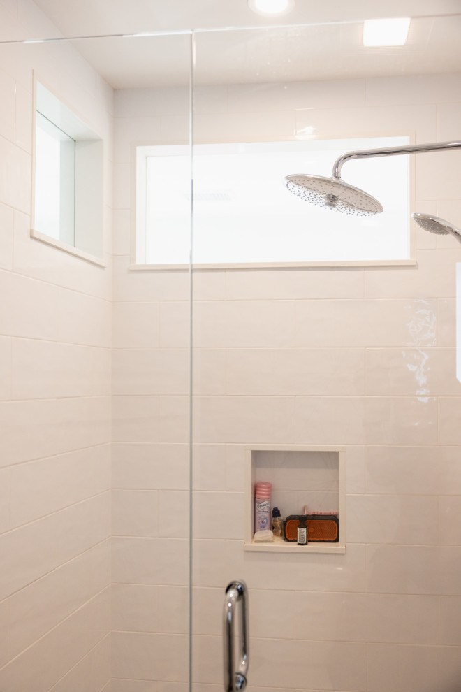 A modern bathroom shower with white tiled walls, a built-in niche holding toiletries, and a dual showerhead system. A glass door with a chrome handle is visible in the foreground. Natural light streams in from a rectangular window above the showerheads.