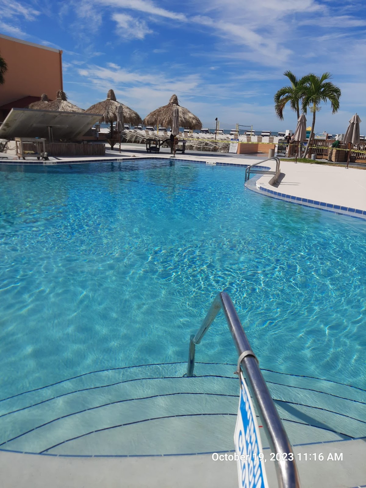 Large resort-style swimming pool with clear blue water, curved entry steps, palm trees, and lounge areas.