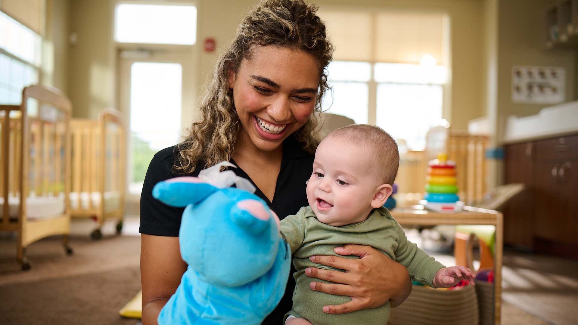 Primrose Schools infant teacher introducing baby to blue stuffed animal puppet in bright infant care room
