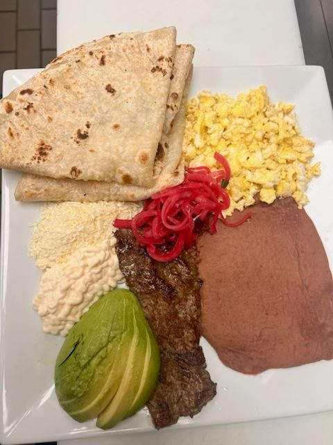 A traditional Honduran breakfast featuring grilled steak, scrambled eggs, refried beans, avocado, queso fresco, crema, and fresh tortillas at Catramex Restaurant.