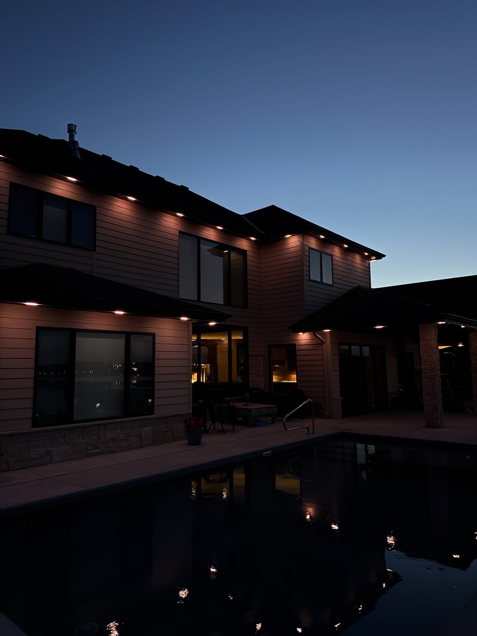 Subtle warm exterior lighting accenting the upper roofline and architectural features of a modern residence beside a pool.
