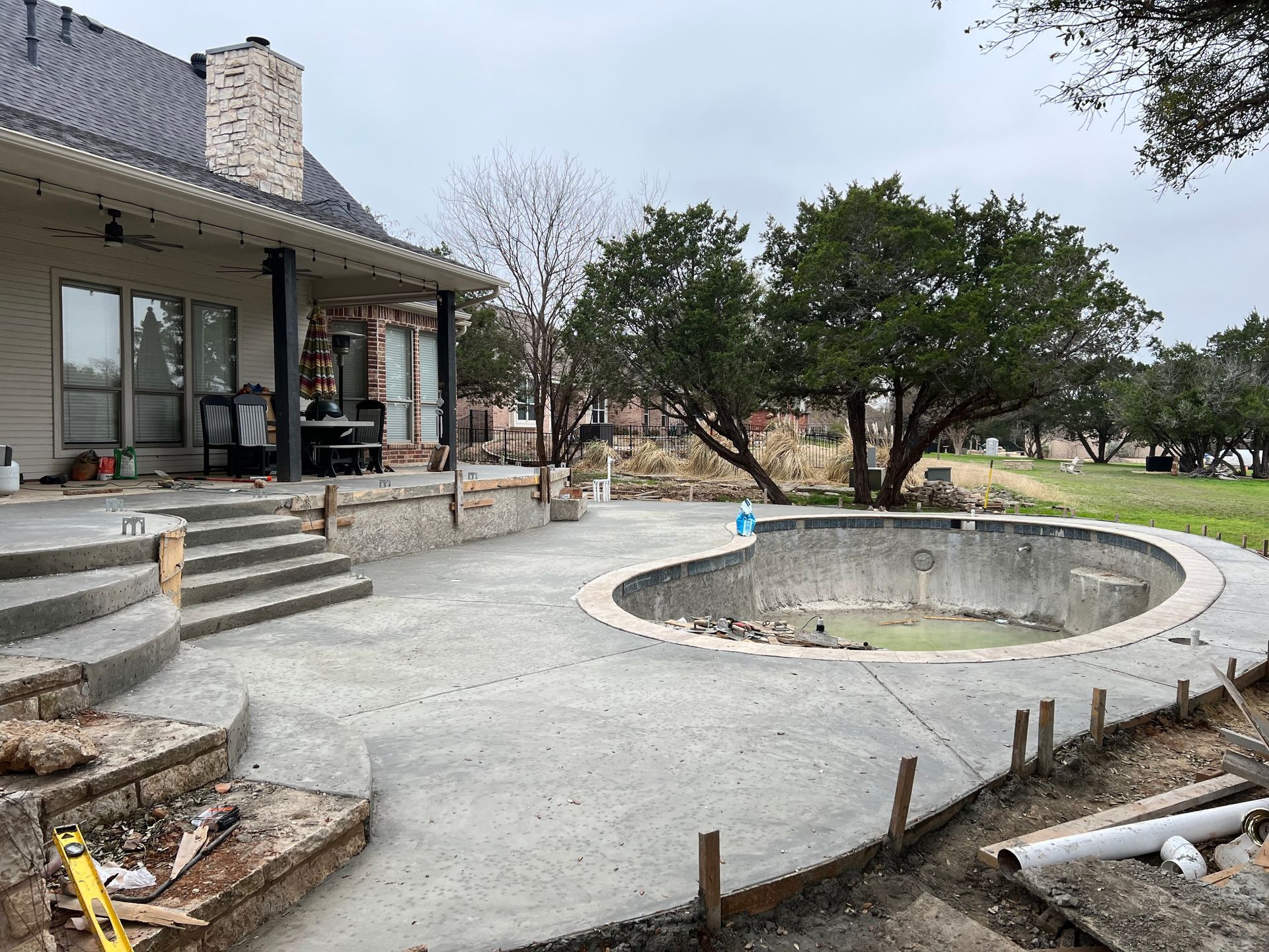 This view showcases a large, multi-level concrete deck with integrated stairs connecting a brick home&rsquo;s patio to a curved pool shell. The construction site is framed by mature trees and greenery.
