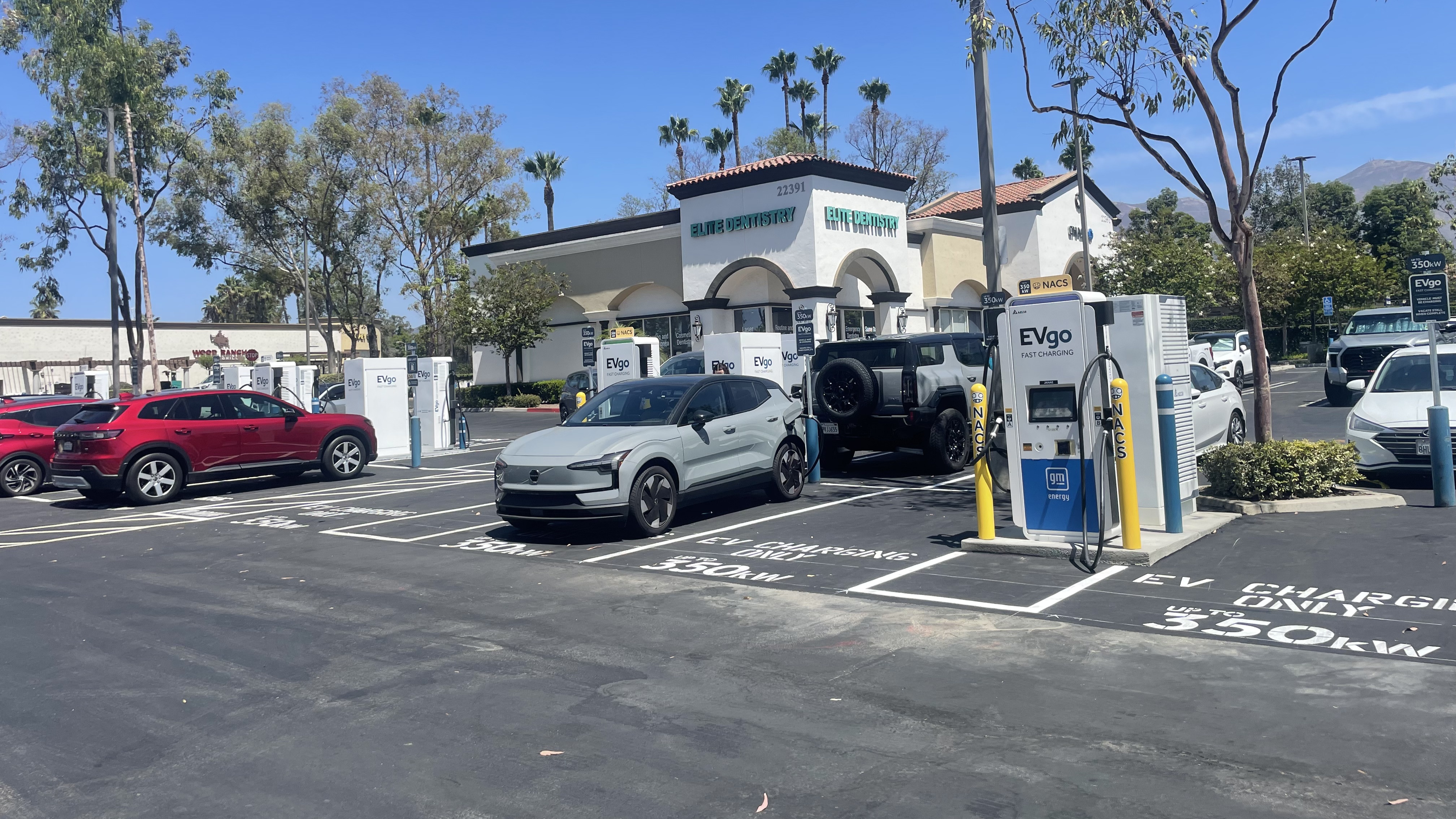 Several EVgo fast charging units installed in marked parking spaces at a public charging station, with EVgo logos on the chargers, protective bollards around each unit. NACS and CCS chargers featured in the image.