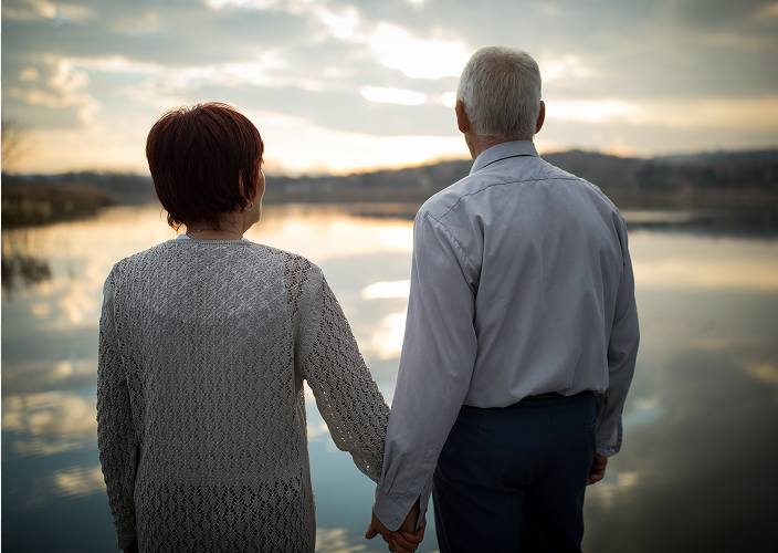 An elderly couple holding hands and looking out towards a lake as sun begins breaking through cloud cover.