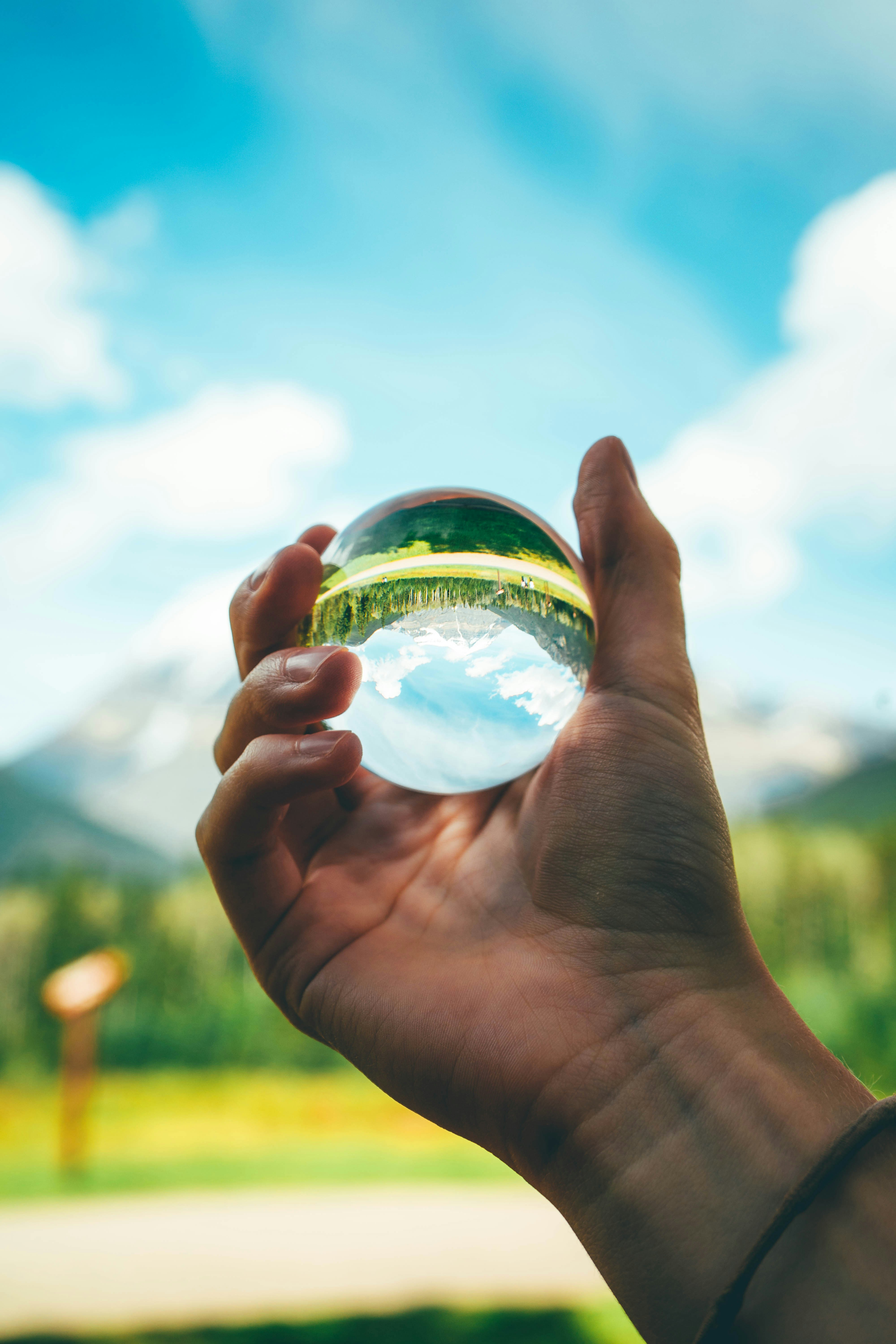 Hand holding clear crystal sphere reflecting inverted landscape against blue sky with clouds