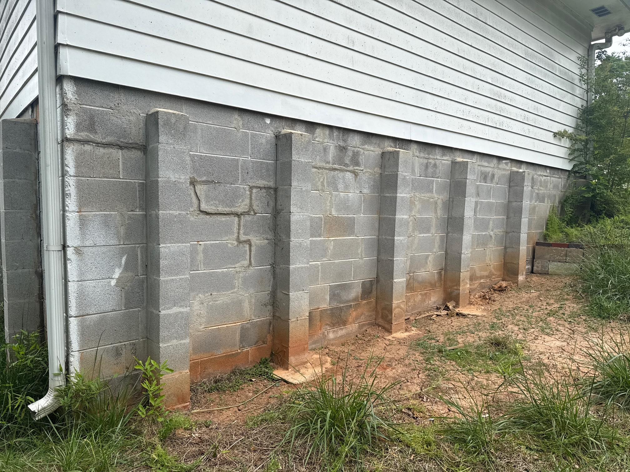 Exterior concrete block foundation wall of a home with visible cracks, staining, and added pilaster reinforcements along the base.