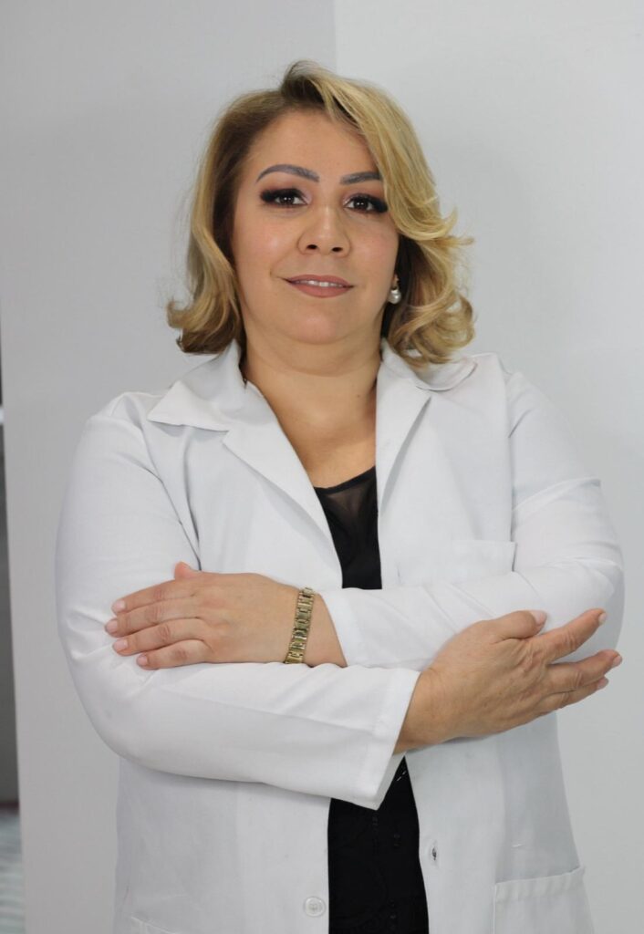 A professional portrait of a woman wearing a white lab coat and a black blouse, with her arms crossed. She has blonde hair and is wearing makeup, including eyeshadow and lipstick. She is also wearing pearl earrings and a gold bracelet.