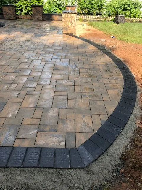 A newly paved patio area with interlocking stone pavers in shades of tan, brown, and gray, bordered by darker charcoal pavers. A stone pillar stands on the left side of the patio. The patio is adjacent to a lawn and garden area with lush green foliage.