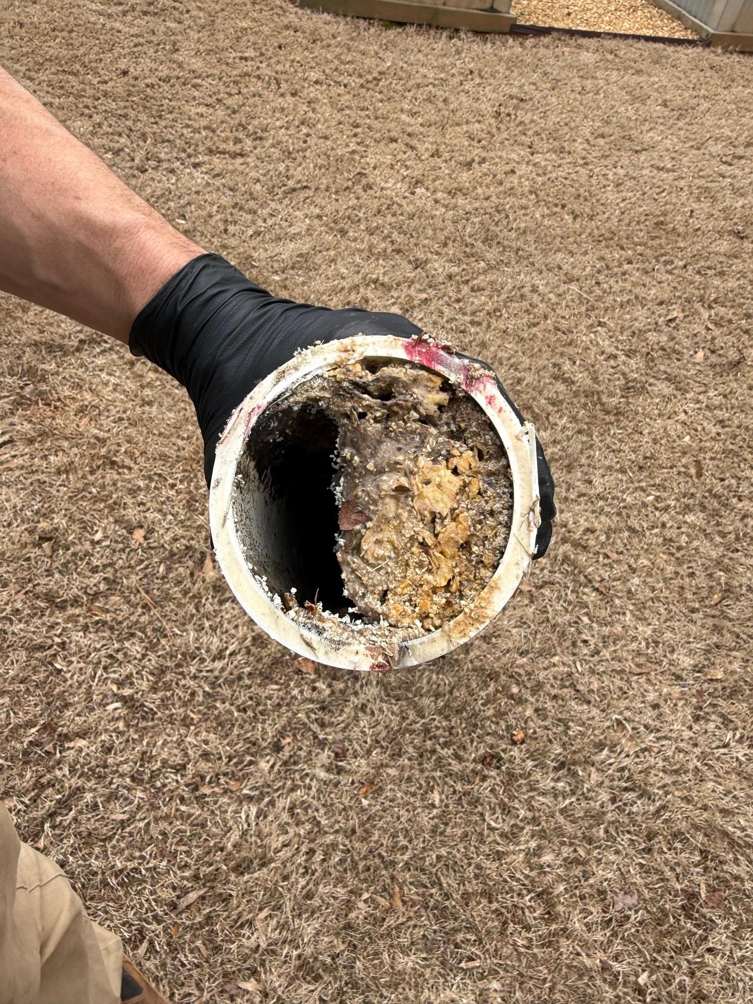 A gloved hand holding a section of white PVC sewer pipe showing heavy buildup and blockage inside the pipe.