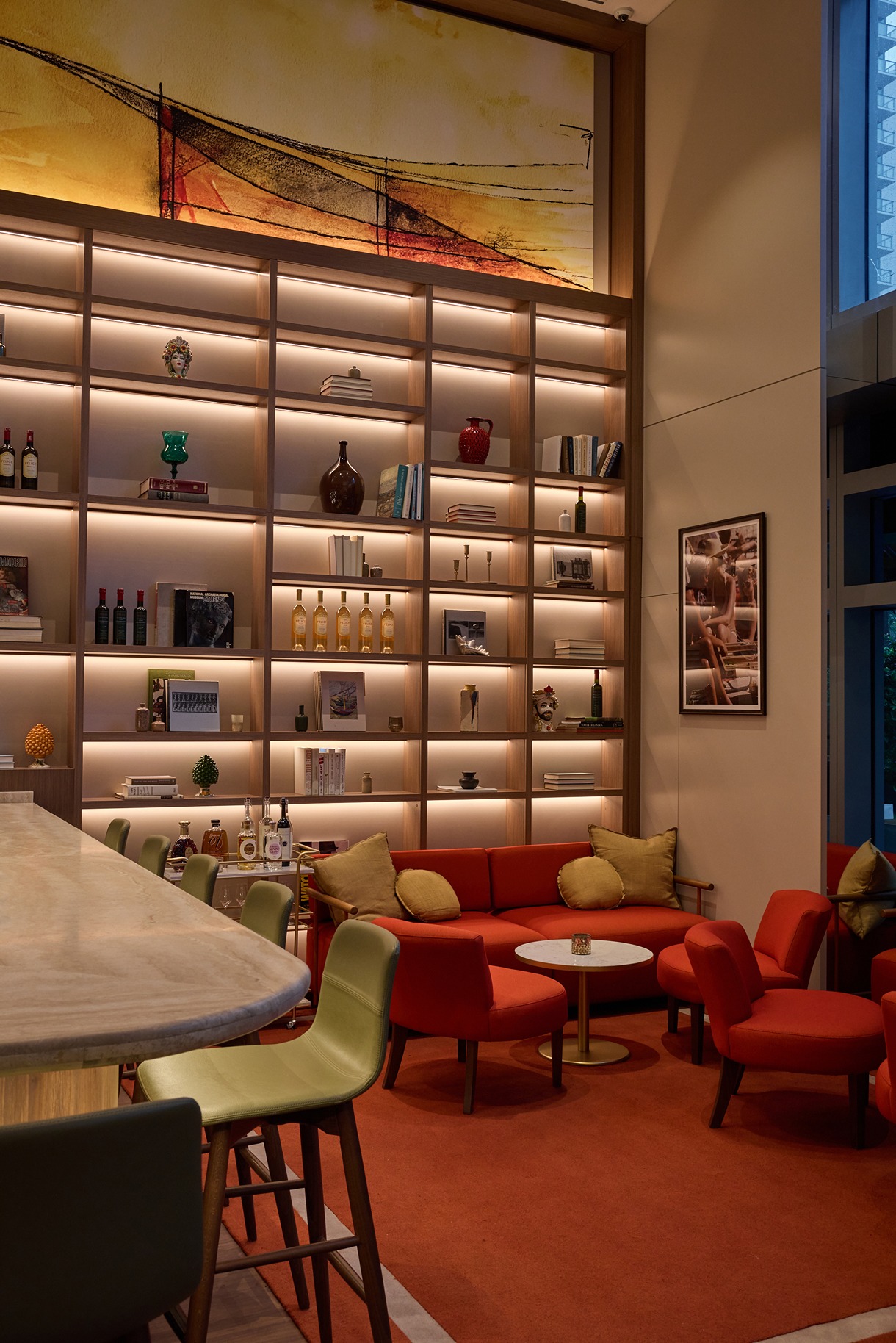 A sophisticated interior with plush red armchairs, an orange rug, and a floor-to-ceiling wooden library wall displaying wine, books, and Italian art pieces.