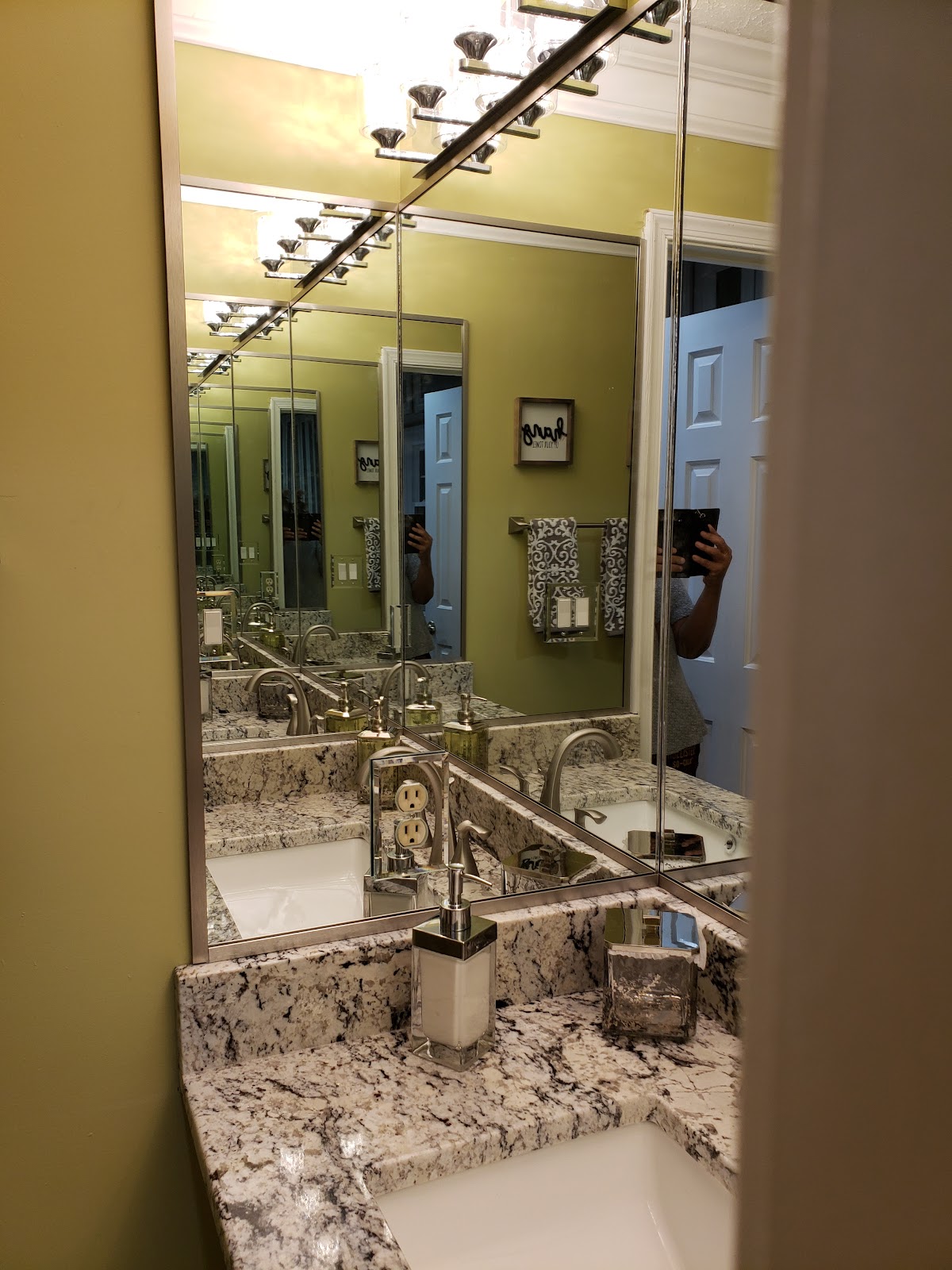 A person is taking a picture of themselves in a bathroom mirror. The bathroom has a double vanity with granite countertops. There are two sinks with chrome faucets. In the reflection of the mirror, we can see a light yellow wall with framed artwork and towels hanging on a rack. There are also lights above the mirror.