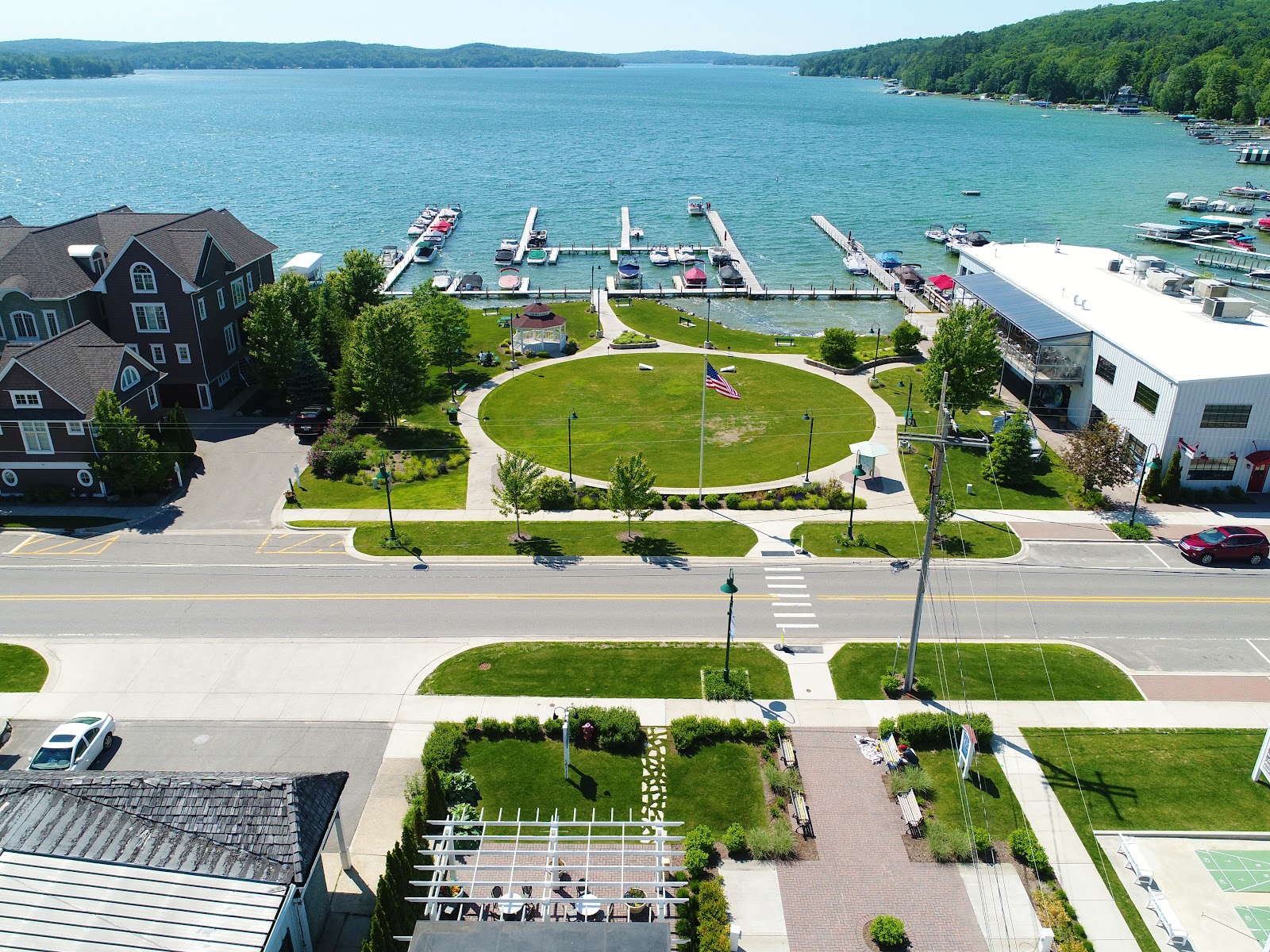 An aerial drone view captures a lakeside town on a clear sunny day. The scene showcases a large, tranquil blue lake dotted with numerous boats moored at wooden docks. Along the shoreline, there are several modern buildings, including a large white building with a dark-paneled roof, and multi-story brown residential buildings. A circular green park with an American flag in the center is situated between the buildings and the lake, with a white gazebo on its edge. A paved road with a pedestrian crossing runs in front of the park, separating it from a landscaped area with benches and a white pergola. The surrounding landscape is lush with green grass and trees, and rolling, tree-covered hills form the backdrop against the horizon.