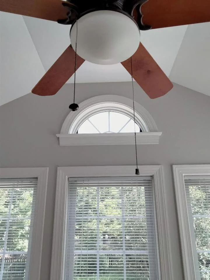 A ceiling fan with wooden blades is shown mounted on a light gray ceiling. Below the fan, a semicircular window with white muntins is visible, framed by white molding. Two rectangular windows with white blinds are also present, showing a view of trees and greenery outside. The image captures a domestic interior with architectural details.