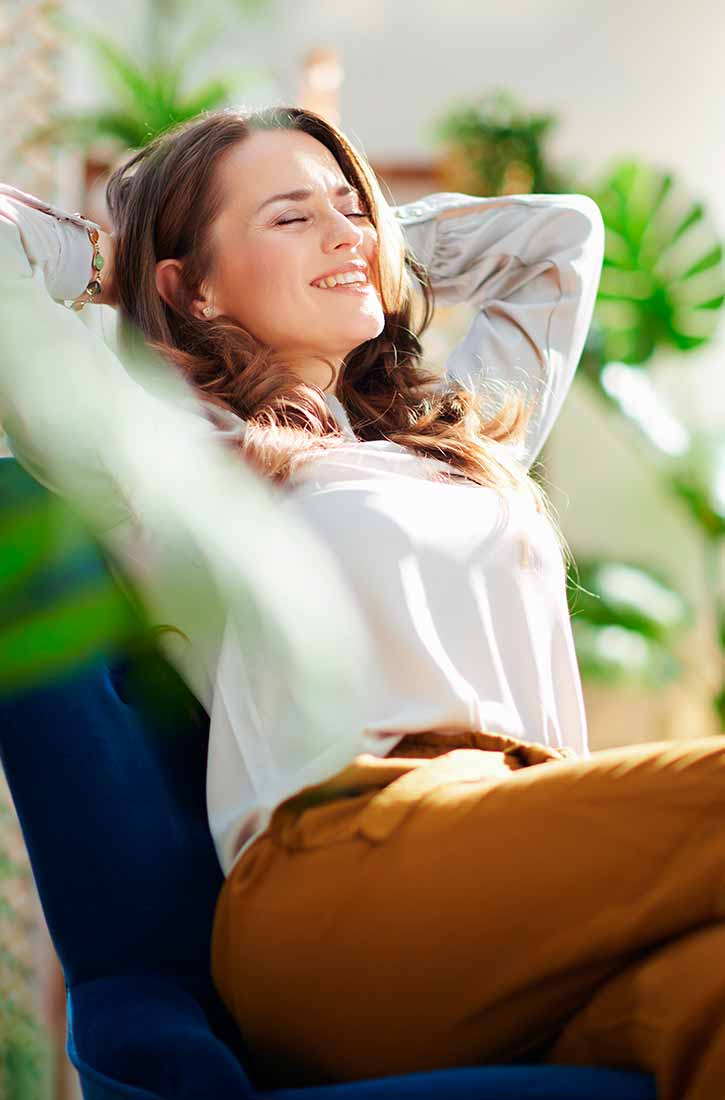 A woman with brown hair and closed eyes smiles as she relaxes in a chair, leaning her head back with her hands behind her head. She is wearing a white blouse and rust-colored pants. Sunlight streams in, illuminating her face and the leaves of plants in the background.