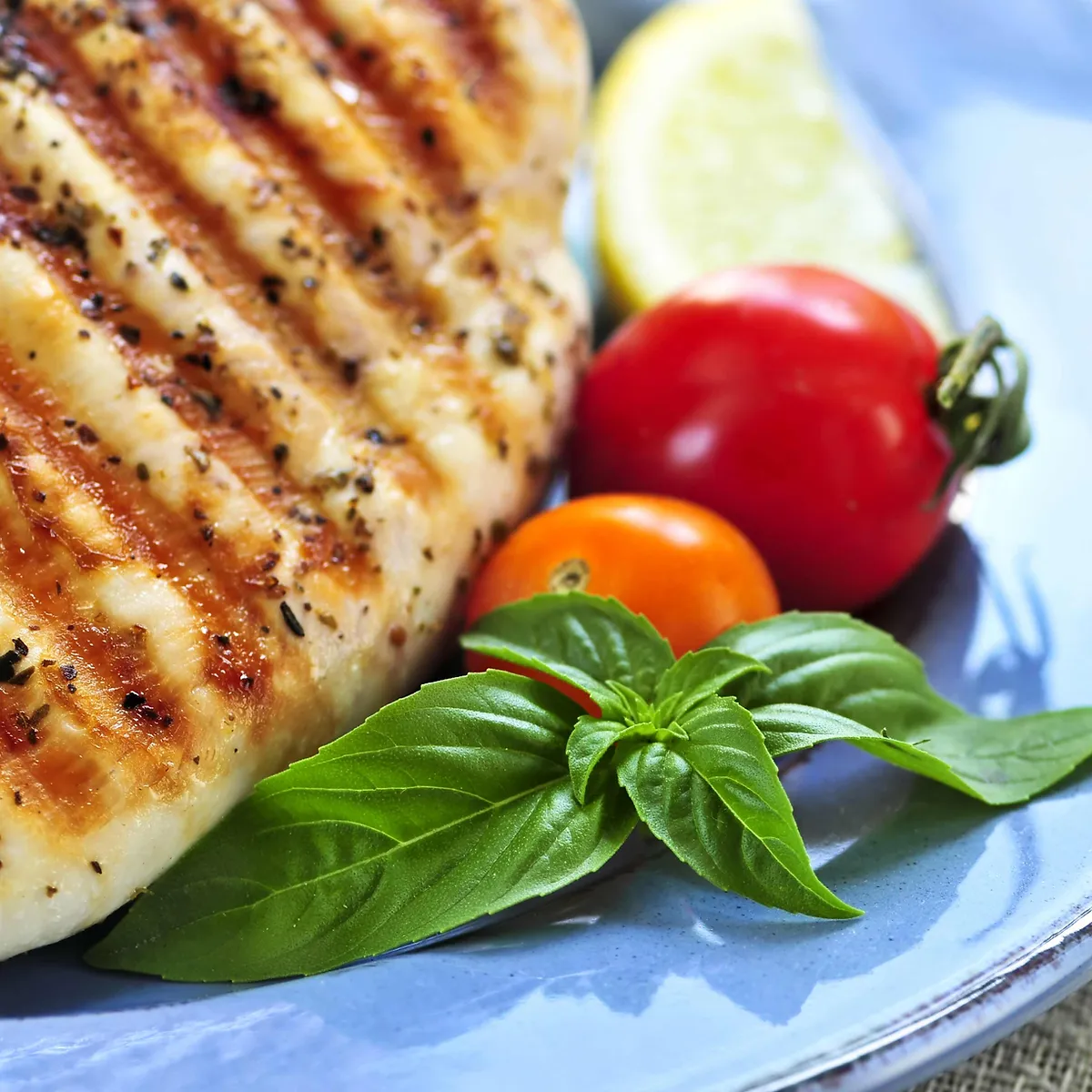 A close-up shot of a perfectly grilled chicken breast seasoned with herbs and spices, served on a blue plate with fresh cherry tomatoes, a slice of lemon, and a sprig of basil.