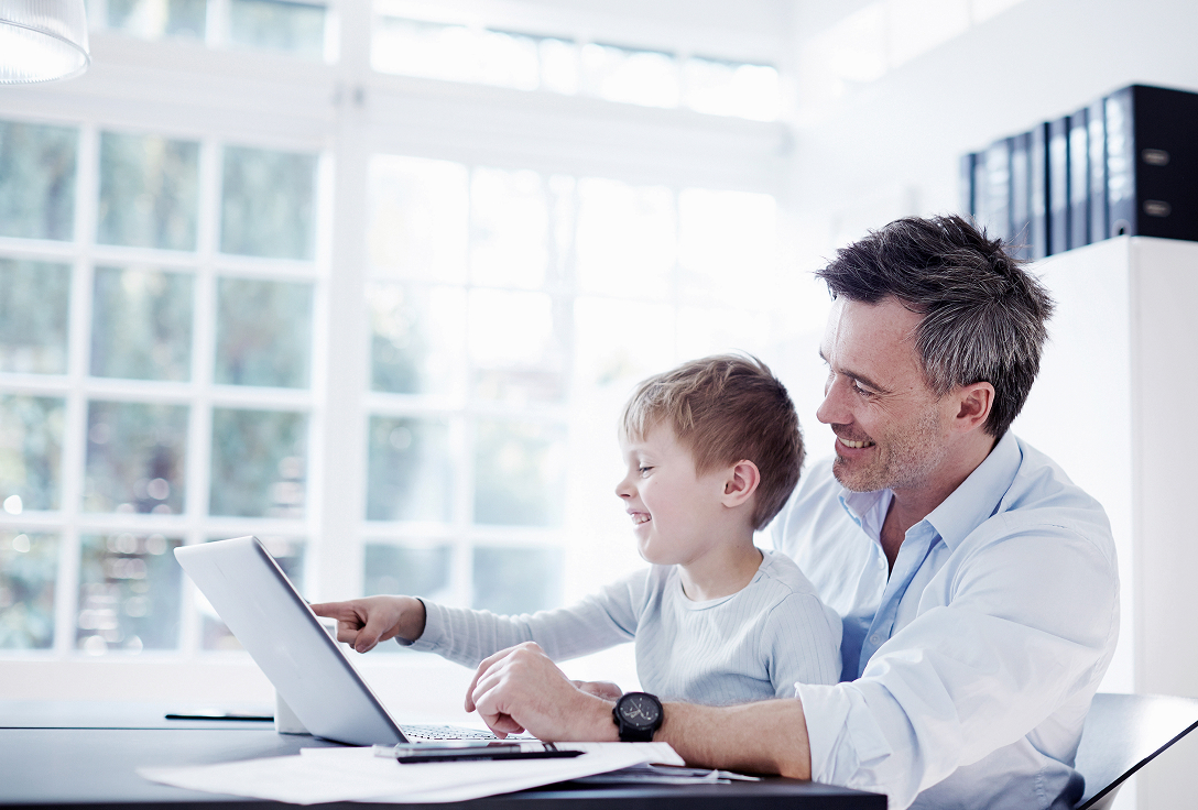 Father and son smiling at a laptop in a bright room with large windows and natural light. The scene conveys happiness and bonding.
