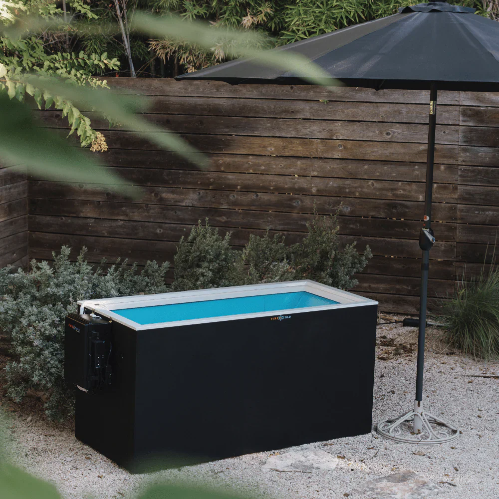 An outdoor cold plunge tub is situated on a gravel patio, surrounded by lush greenery and a rustic wooden fence. A large black umbrella stands nearby, providing shade. The tub is filled with vibrant blue water and features a control unit attached to its side. The branding "FIRE & COLD" is visible on the tub.