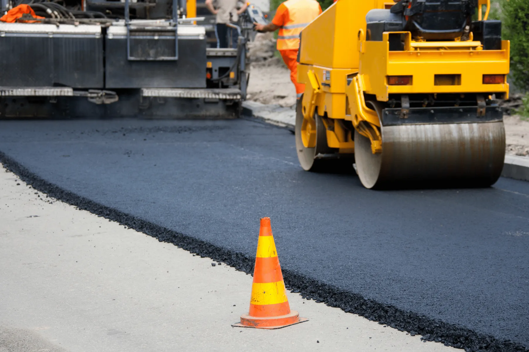A bright orange and yellow traffic cone stands in the foreground on a gray surface next to a freshly laid strip of black asphalt. In the background, heavy construction equipment, including a large yellow road roller and a black asphalt paving machine, are actively working on resurfacing a road. Two construction workers are visible, one wearing an orange vest and the other a beige shirt, operating the machinery. The scene depicts an active road construction site.