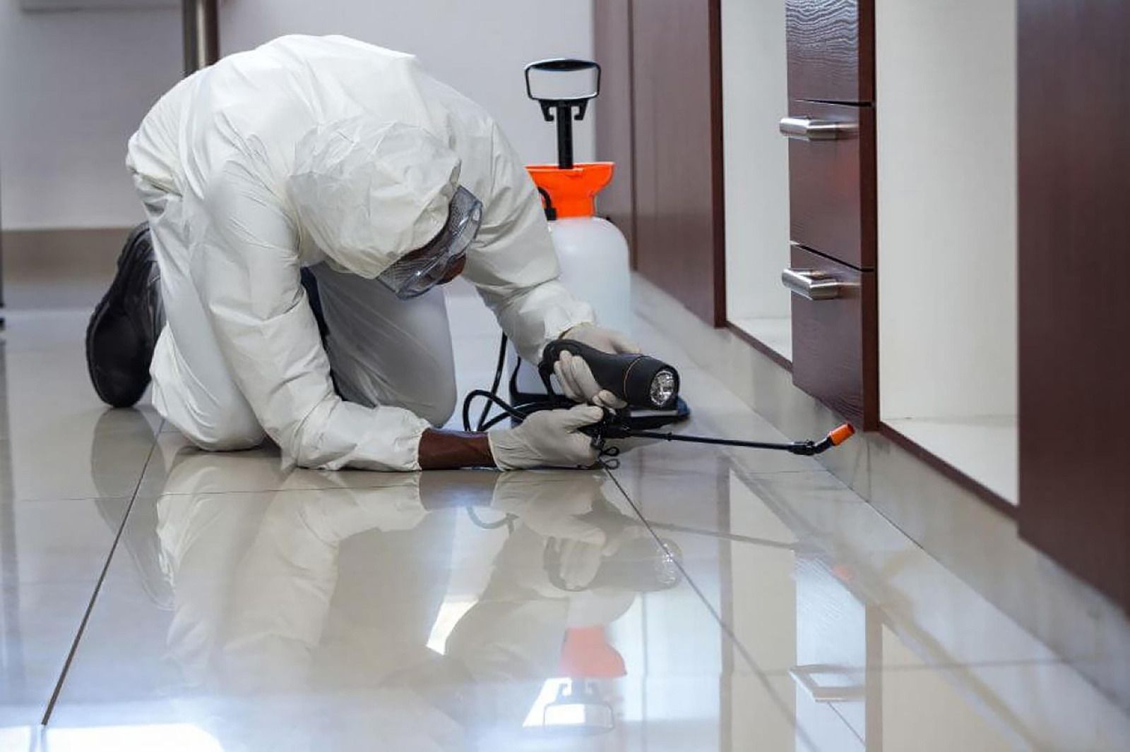 A pest control technician wearing protective clothing kneels on a floor while applying pesticide along the base of kitchen cabinets, targeting hidden pests and preventing infestations in a residential indoor environment.