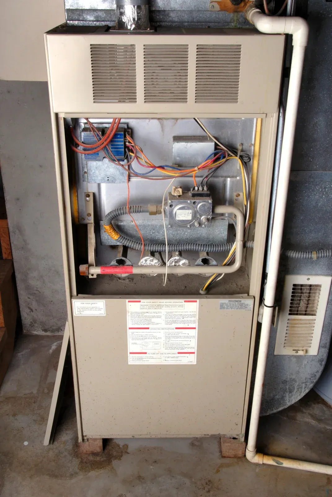 An older residential furnace with its front panel removed, exposing internal wiring, gas components, and blower system, installed in a garage area with visible wear and surrounding concrete flooring.